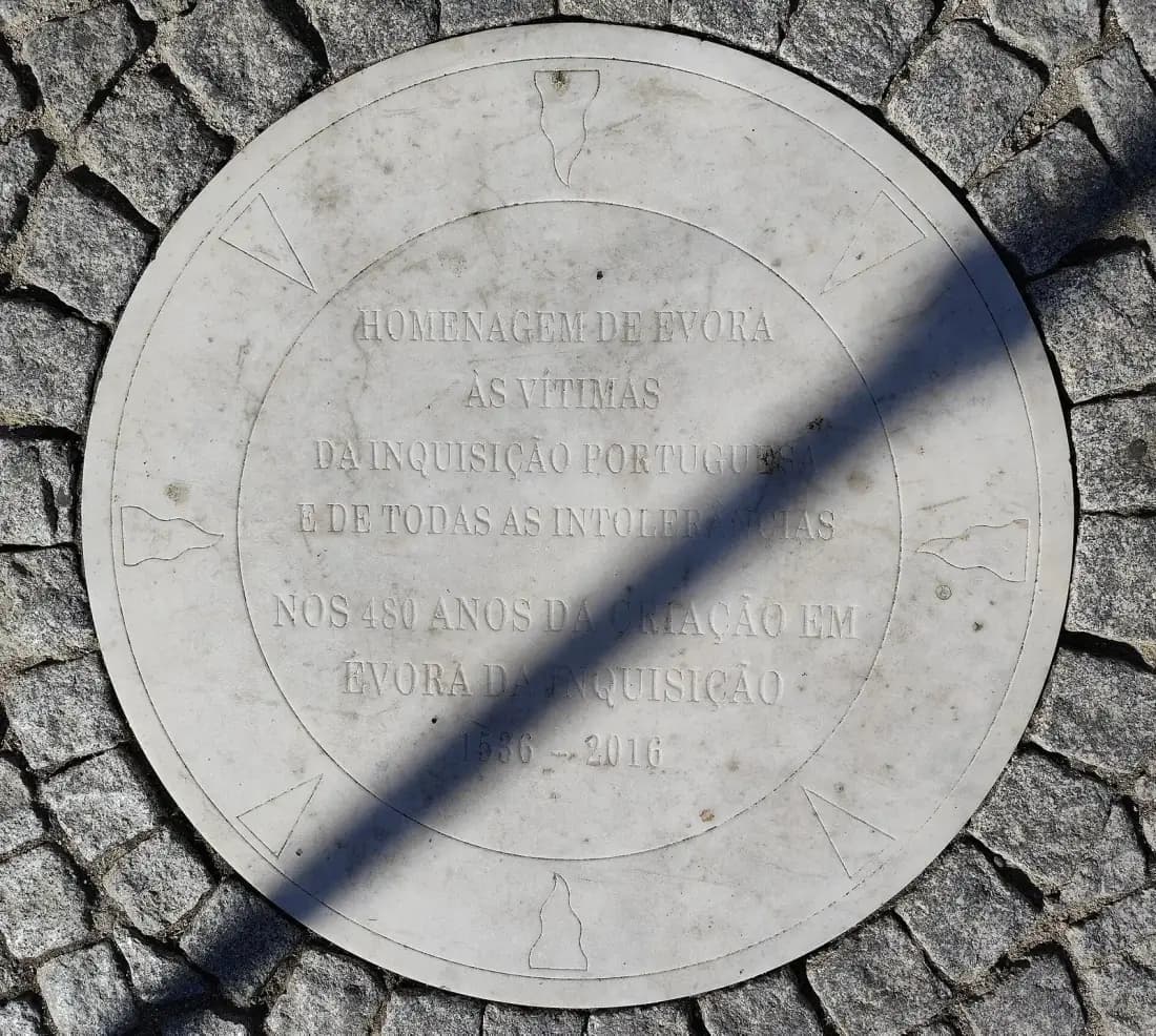 A round white marble plaque set into grey cobblestones, carved with small flame shapes around its rim and an inscription in capital letters in Portuguese; a diagonal band of shadow crosses it.