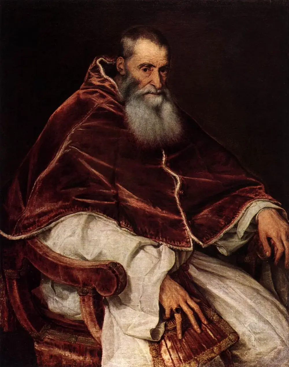 Painted portrait of an elderly, balding man with a long grey beard, seated in a red velvet armchair against a dark background. He wears a heavy dark-red velvet cape over a white robe, and one ringed hand rests on a purse in his lap.