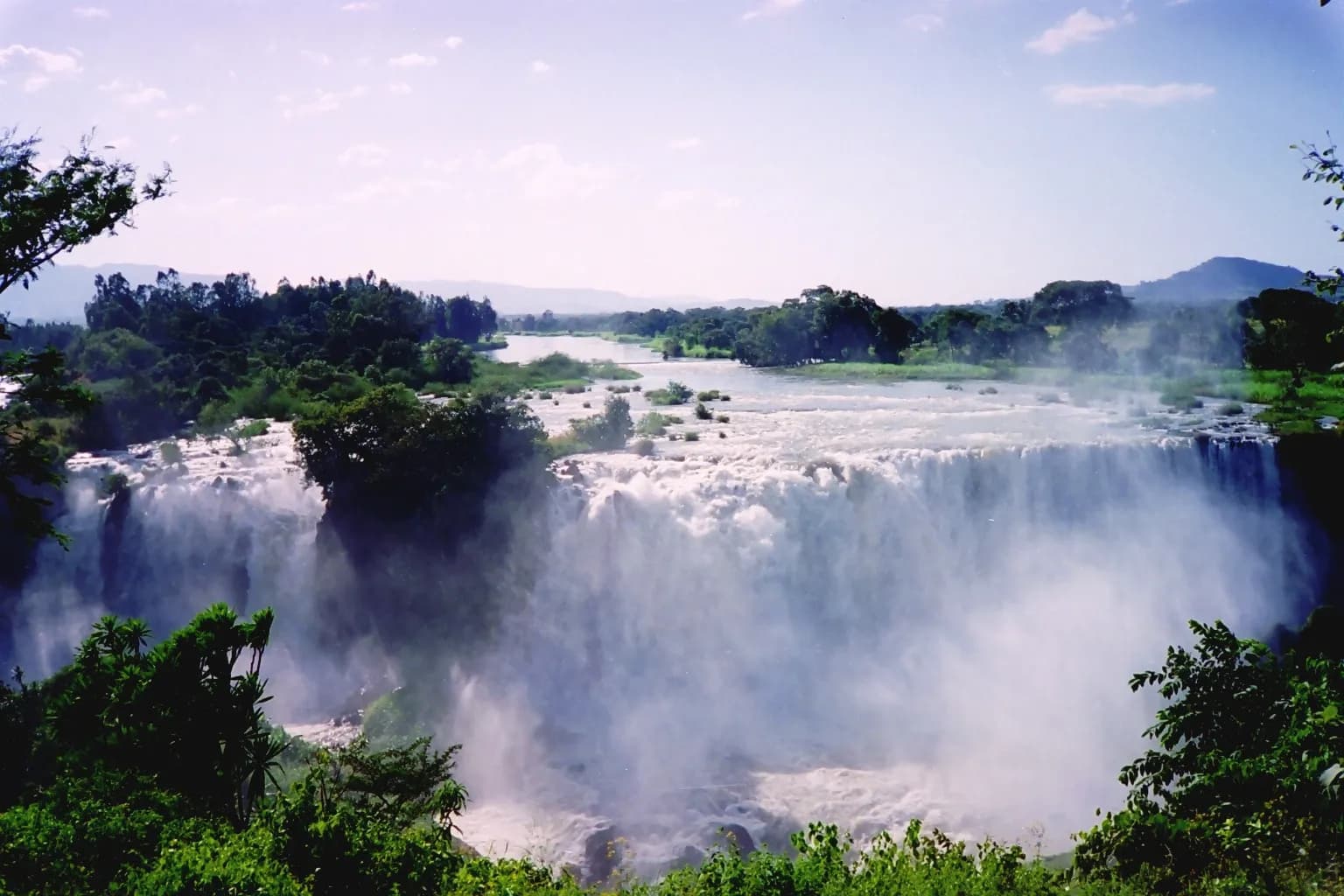 A broad waterfall pours over a rock ledge in several white curtains, throwing up clouds of spray, with green trees in the foreground and a wide river and hills behind.