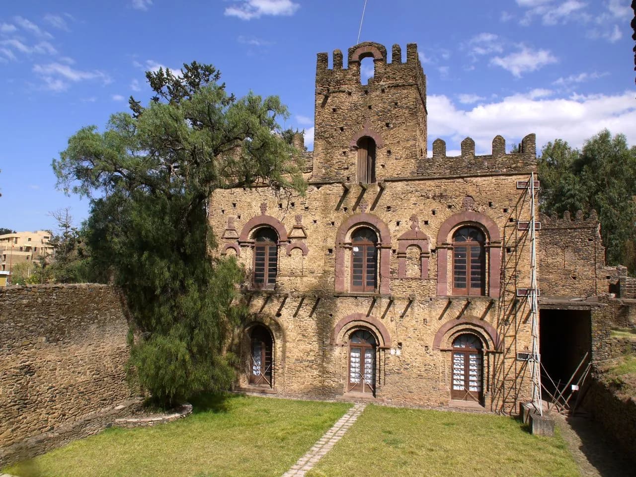 A tall stone building with crenellated walls, a square battlemented tower and rows of arched windows trimmed in reddish stone, beside a lawn and a large tree under a blue sky.
