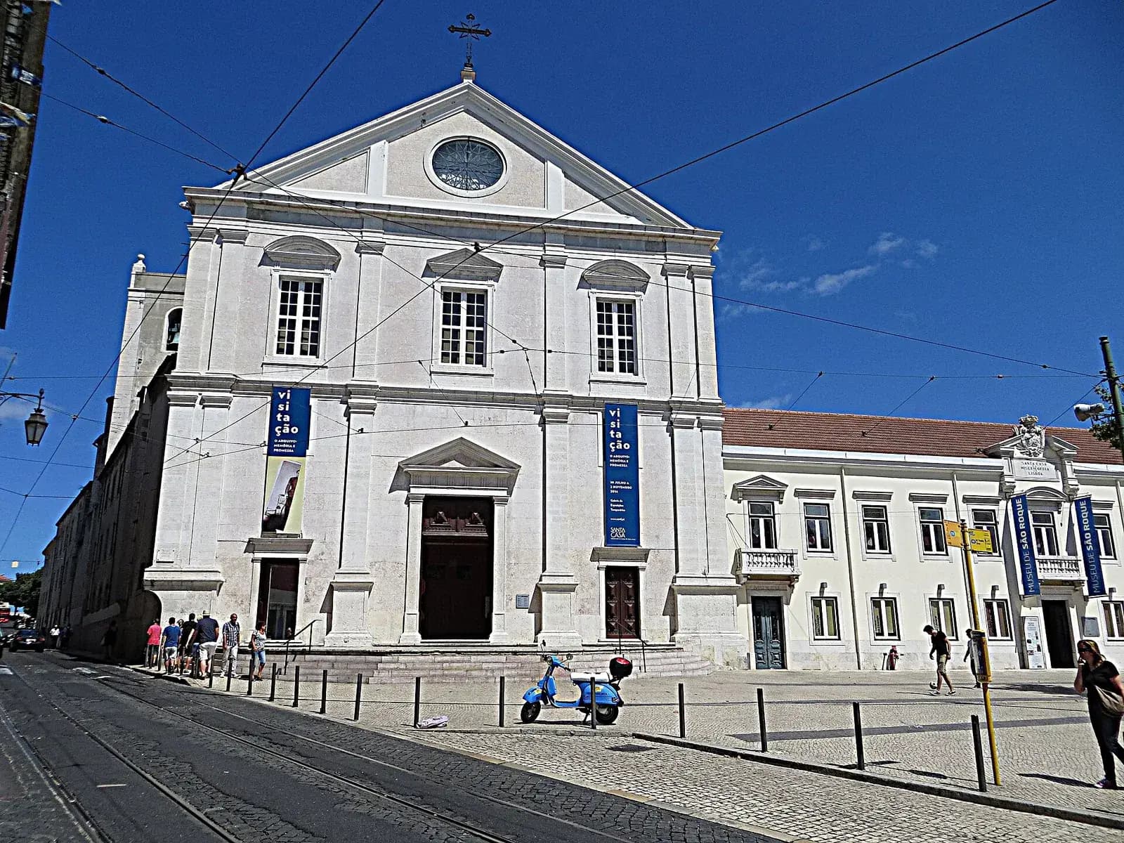 The plain white stone front of a church with a triangular pediment, a round window and three tall windows above the doors, hung with blue banners. To its right is a long, low white building with blue Museu de São Roque banners, and a scooter stands on the cobbled square in front under a deep blue sky.
