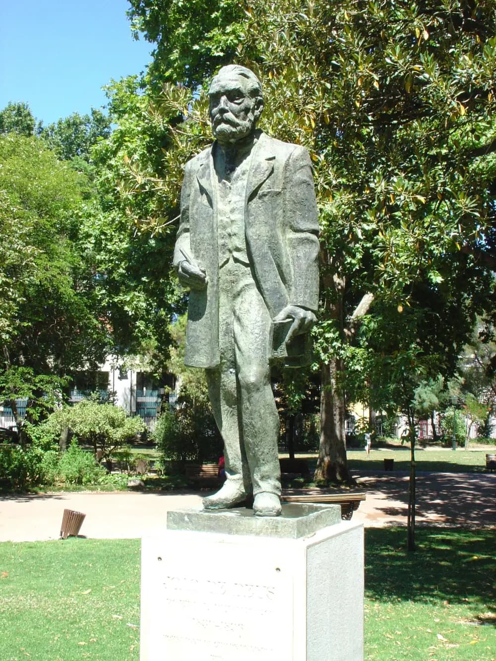 A dark bronze statue of an elderly bearded man in a long coat, holding a pen in one hand and a book in the other, stands on a pale stone plinth on a lawn under tall trees.