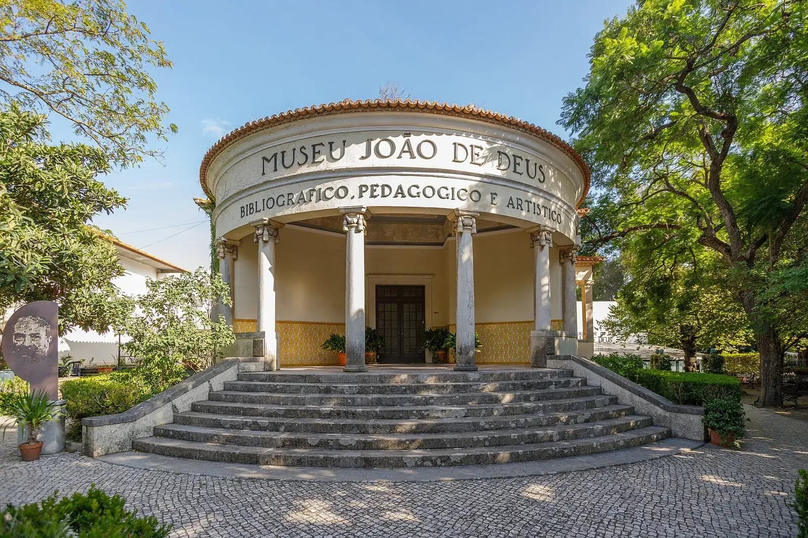 A round, single-storey pavilion with a red tiled roof and a ring of white columns, reached by a wide flight of stone steps. Lettering above the columns reads Museu João de Deus. Trees stand on either side.