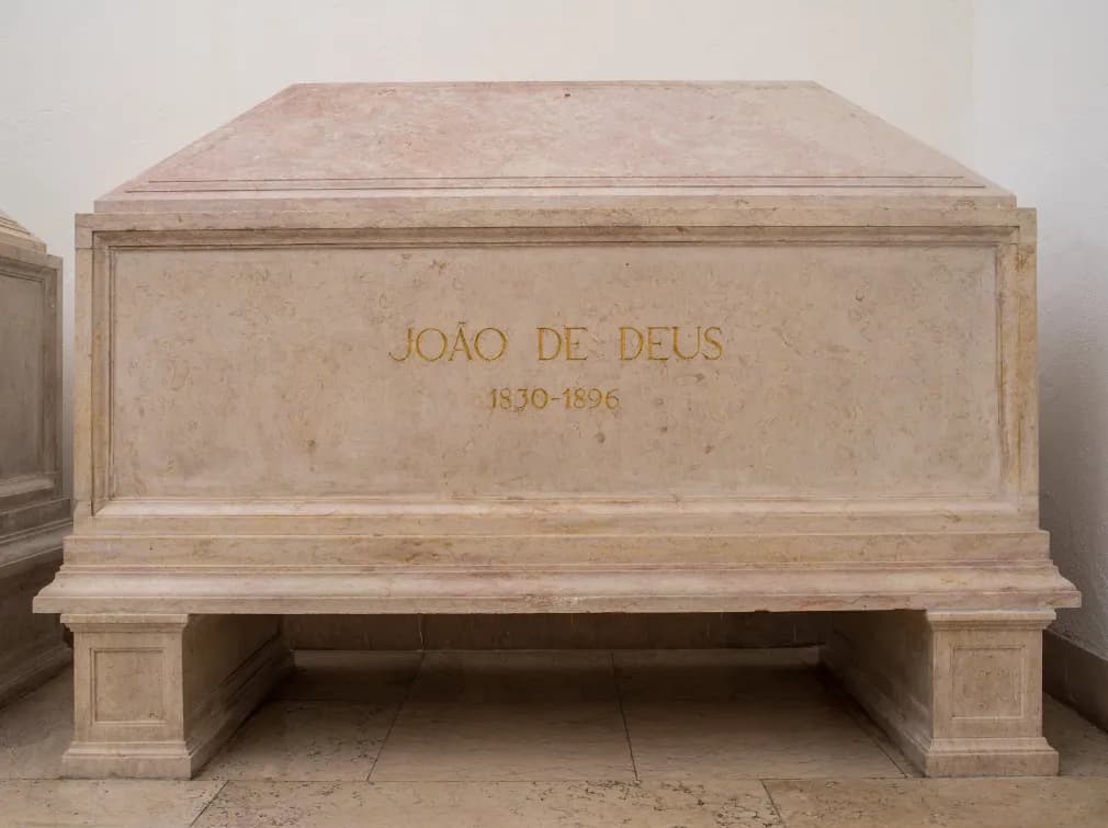 A plain pinkish marble tomb on a low base against a white wall, carved with the words João de Deus 1830-1896 in gold letters.