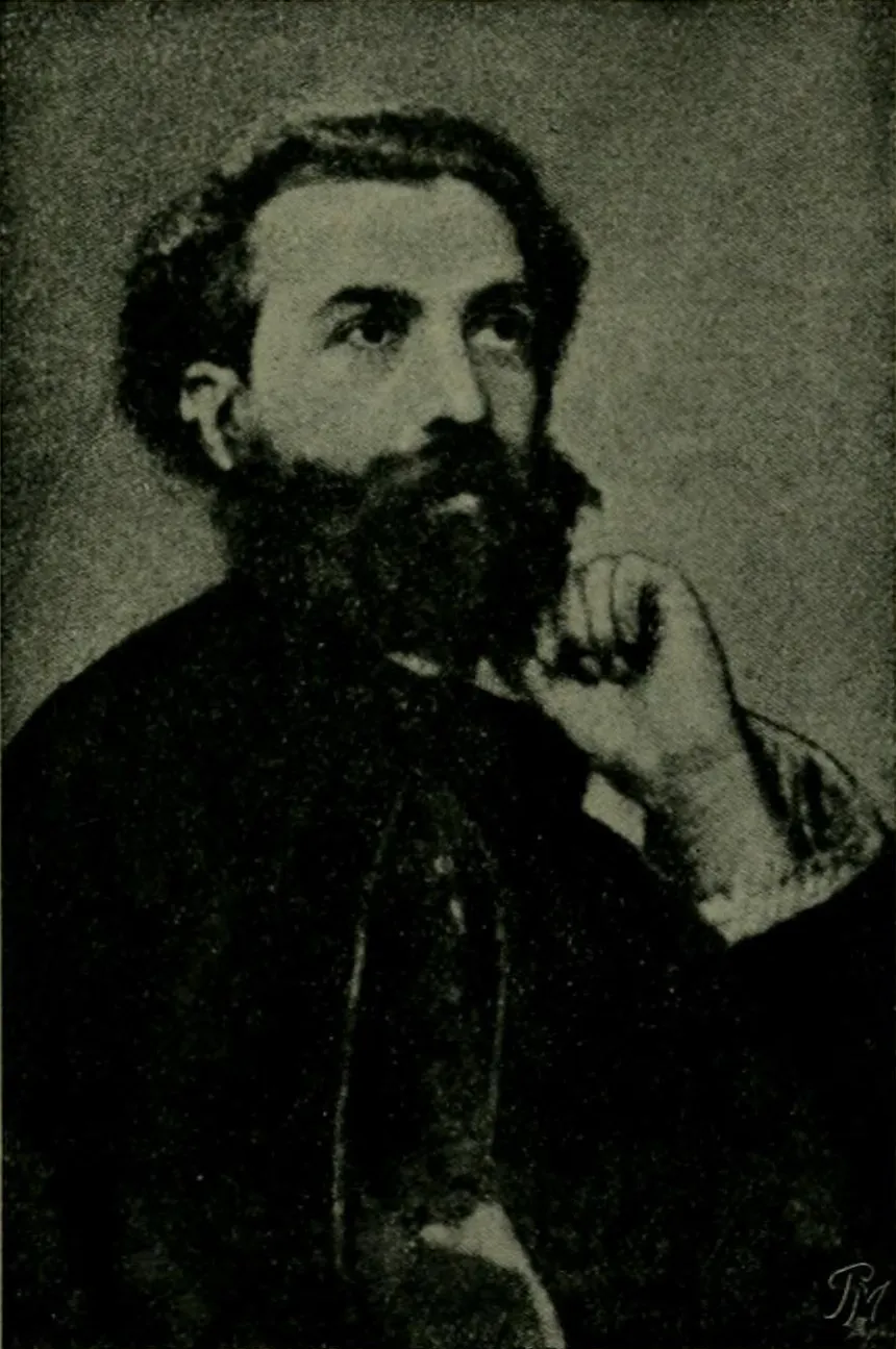 A young man with thick dark hair and a full beard sits with his hand raised to his chin, looking up and to one side, in a dark jacket.
