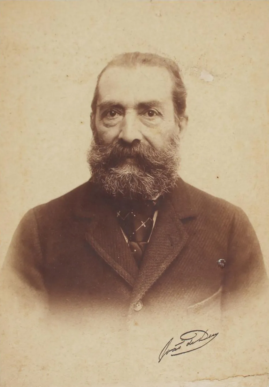 Sepia studio photograph of an older man with a long greying beard and a high forehead, wearing a dark jacket and tie, looking straight at the camera. A signature is written across the lower corner.