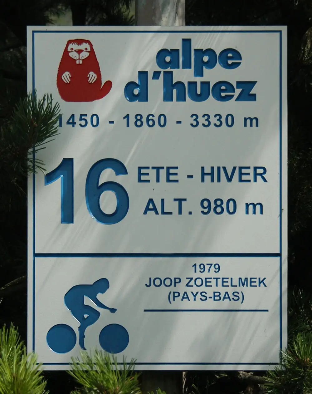 A white roadside sign with a red marmot logo, reading alpe d'huez, 1450 - 1860 - 3330 m, 16, Ete - Hiver, Alt. 980 m, and below a blue cyclist pictogram the words 1979 Joop Zoetelmek (Pays-Bas), with the name misspelt.