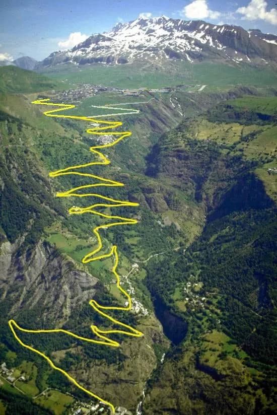 A steep green mountainside seen from above, with the zigzagging road up to Alpe d'Huez traced in yellow from the valley floor to the top, and snow-capped peaks behind.