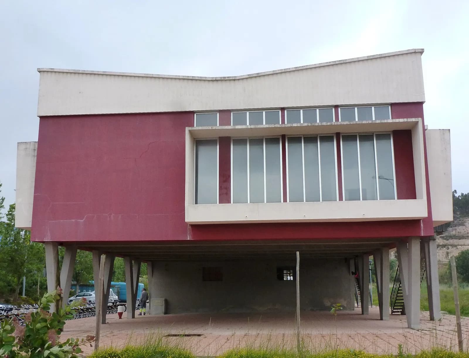 A two-storey modernist building with a dark red and white upper floor that juts out over slender concrete legs, above an open ground floor, against a pale sky.