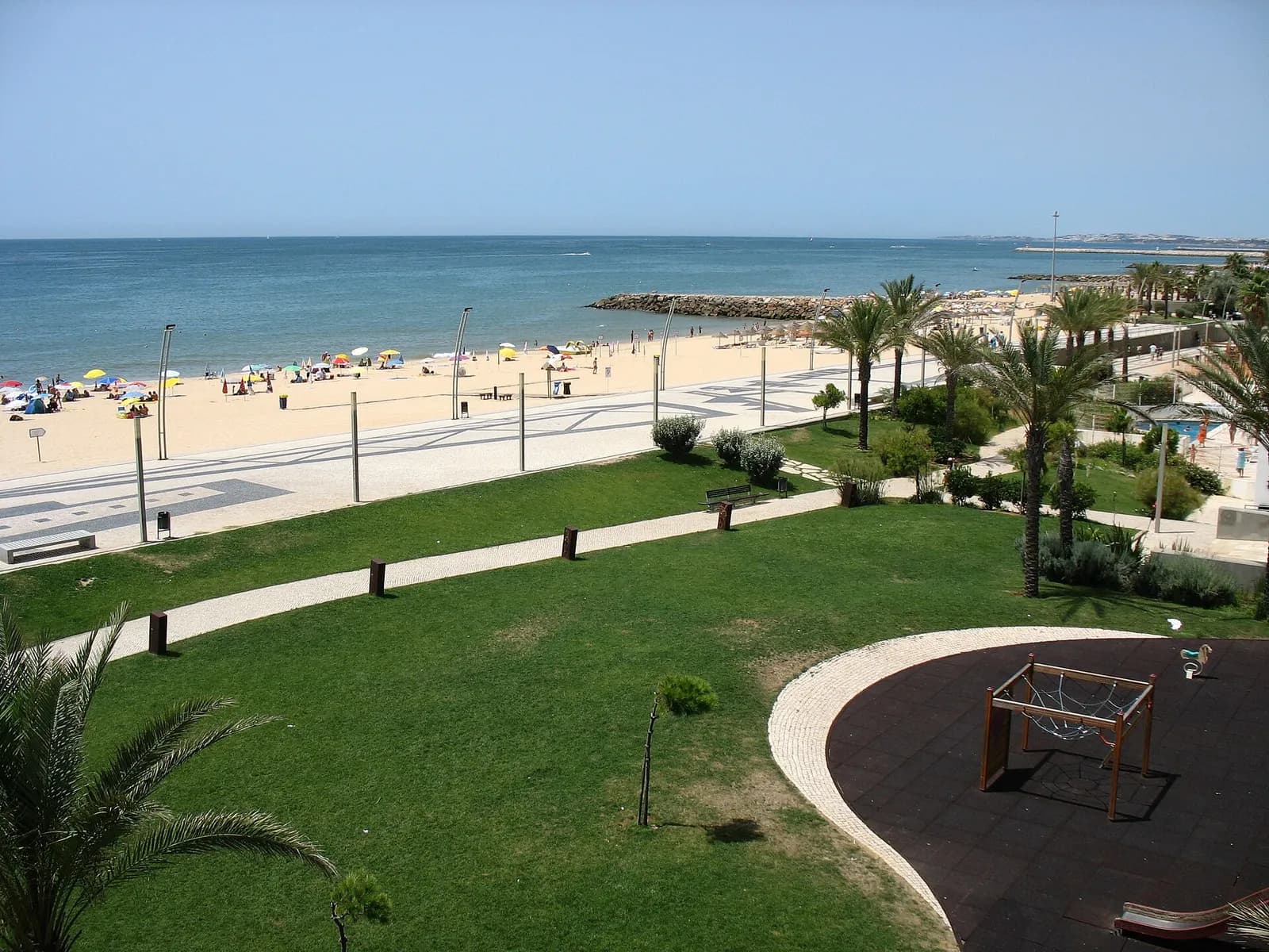 A wide seafront promenade with palm trees, lawns and a playground beside a long sandy beach, with a calm blue sea and a breakwater in the distance.
