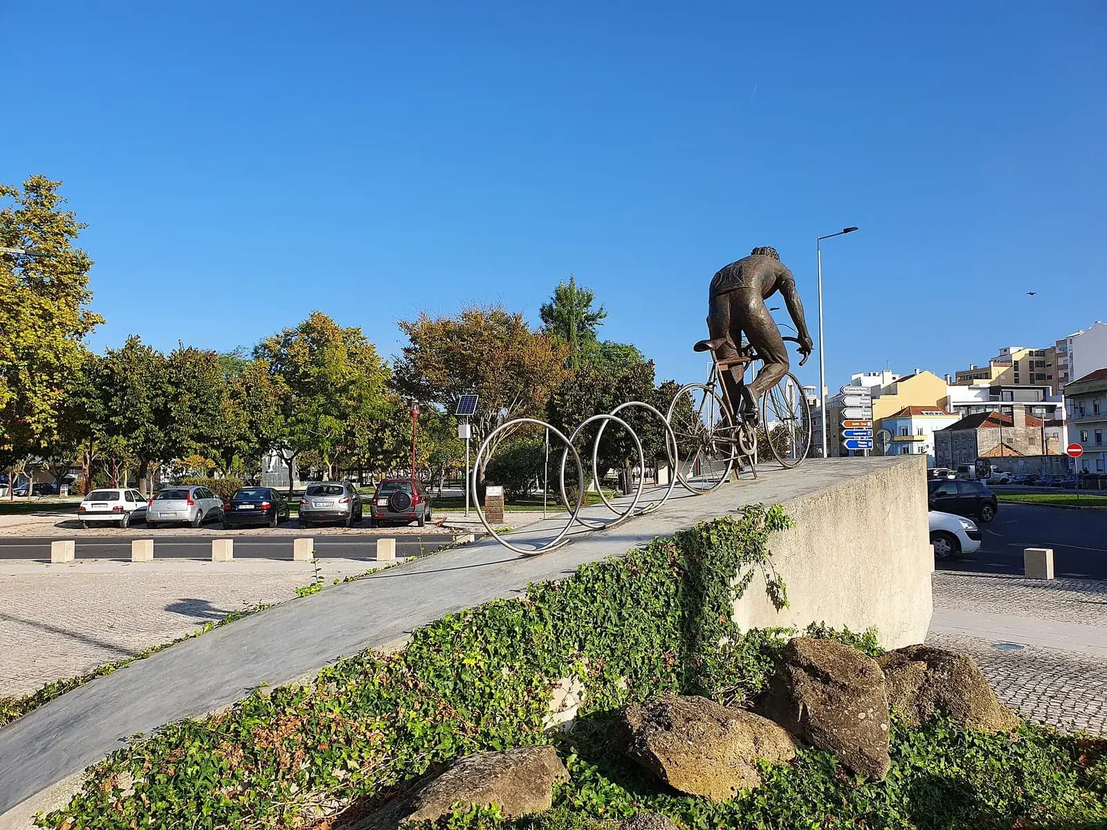 A bronze statue of a racing cyclist, seen from behind, riding up to the top of a sloping concrete ramp. Large metal hoops like bicycle wheels stand in a row behind him, under a clear blue sky, with parked cars, trees and apartment blocks beyond.