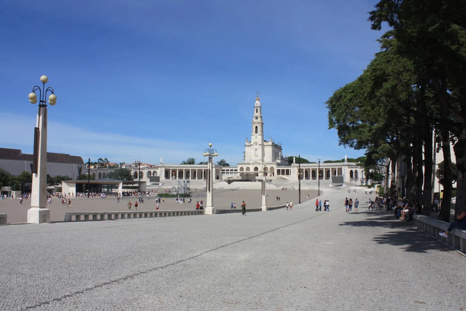 A wide paved square with tall lamp posts and scattered visitors. At its far end a white basilica with a tall bell tower stands at the top of broad steps, flanked by curved colonnades, under a blue sky.