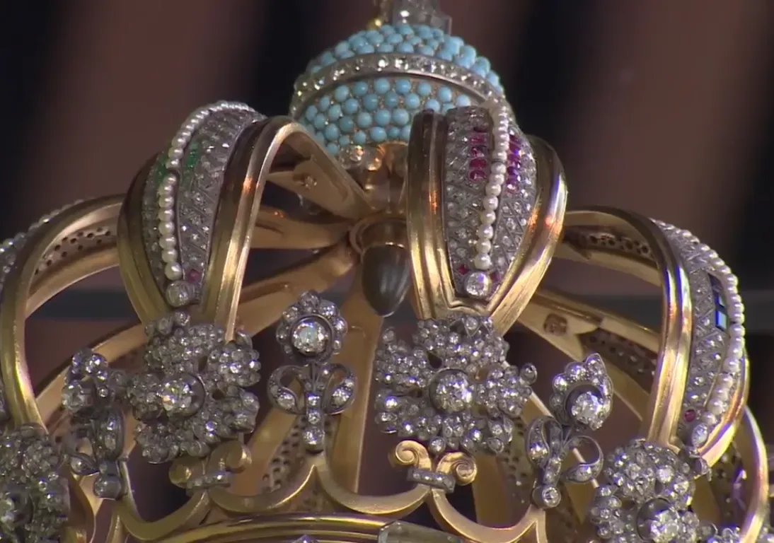 Close-up of a gold crown encrusted with diamonds, pearls, rubies and sapphires. Its arches meet beneath a dome of small turquoise stones, and in the centre, just under the dome, hangs a small dull grey bullet.