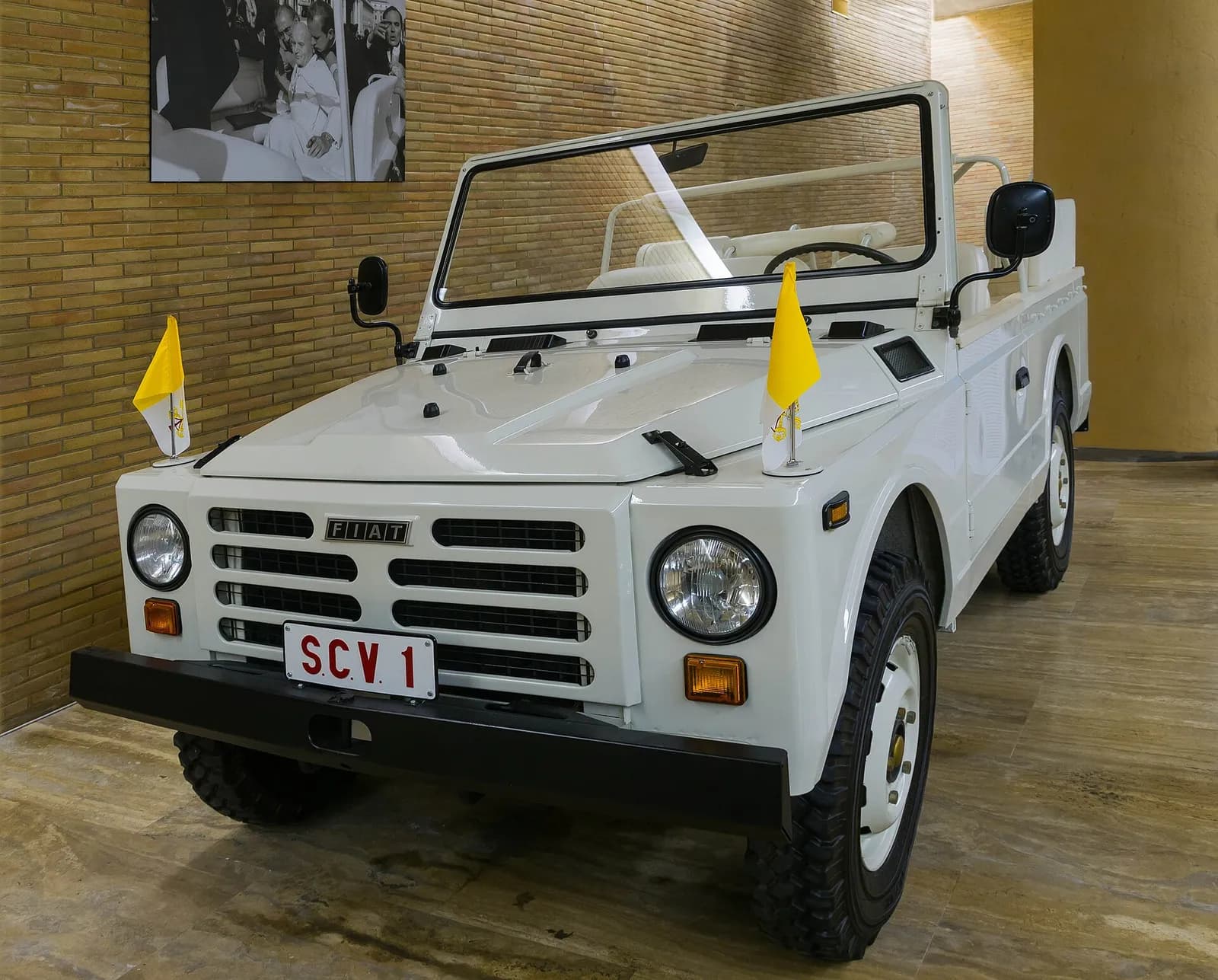 A white open-topped Fiat jeep with a windscreen, a raised rear platform and small yellow and white Vatican flags on the front wings, displayed indoors in front of a brick wall hung with a black-and-white photograph. Its number plate reads SCV 1.