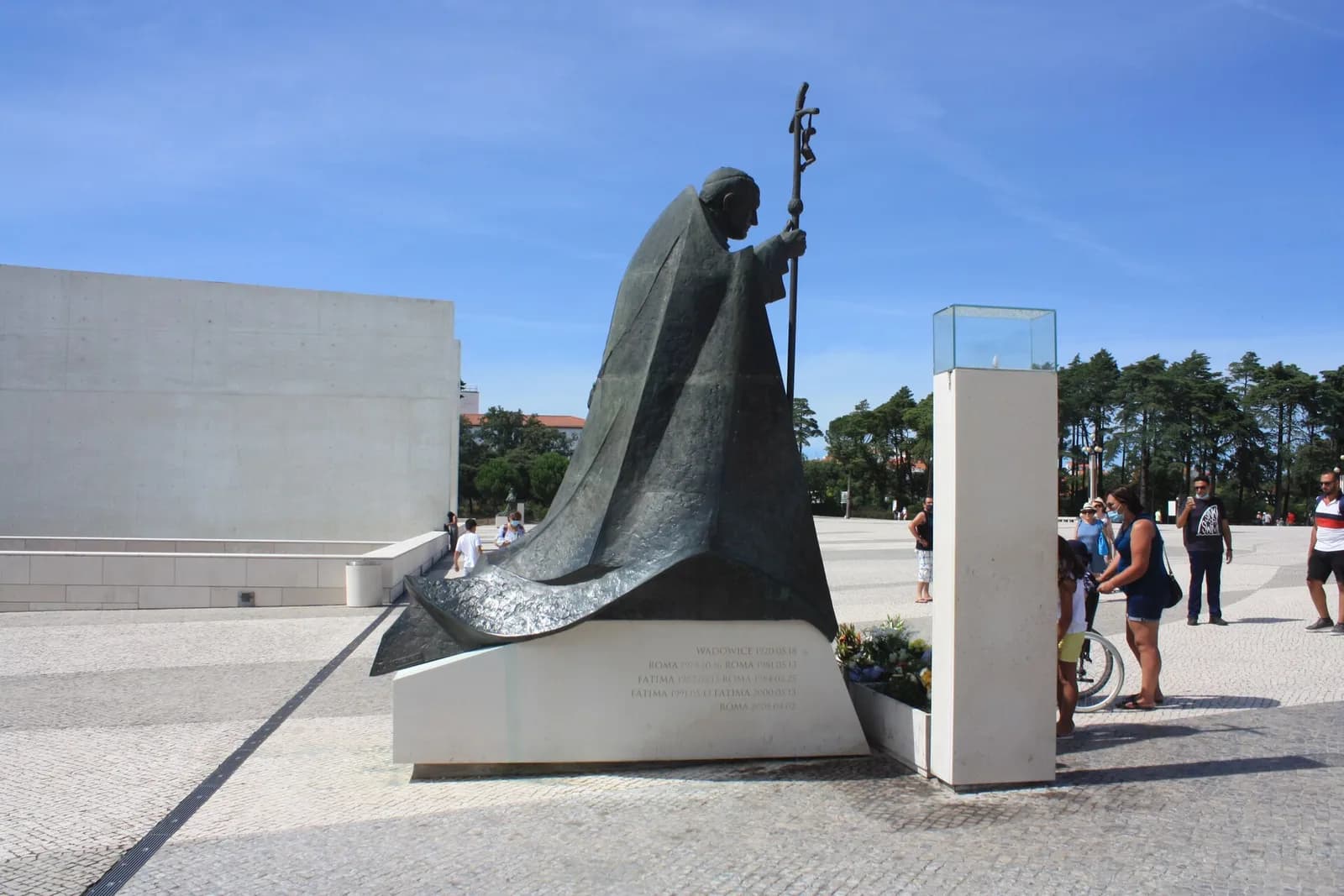 A large dark bronze statue of John Paul II, bent forward under a sweeping cloak and leaning on a tall crucifix staff, stands on a white stone plinth in a paved square, with flowers at its foot and visitors nearby.