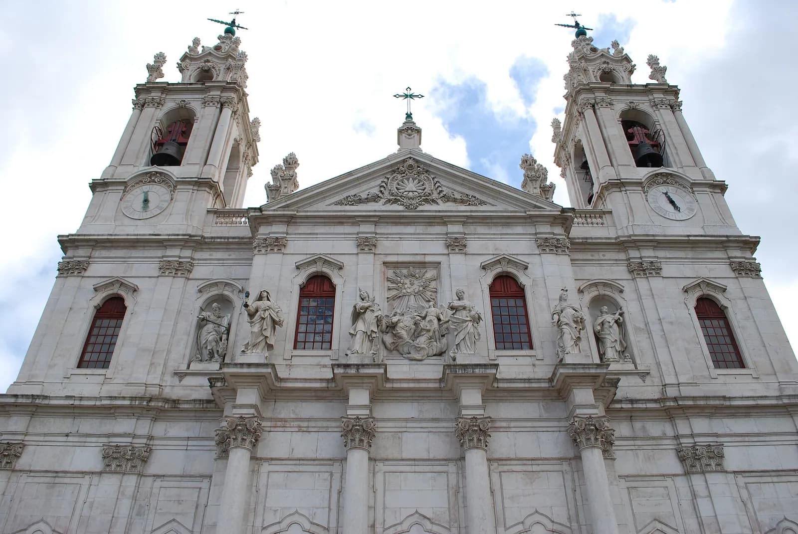 The white limestone front of a large baroque church seen from below, with two bell towers with clocks and weather vanes, a triangular pediment carved with a sunburst, statues of saints and arched red-framed windows between columns.