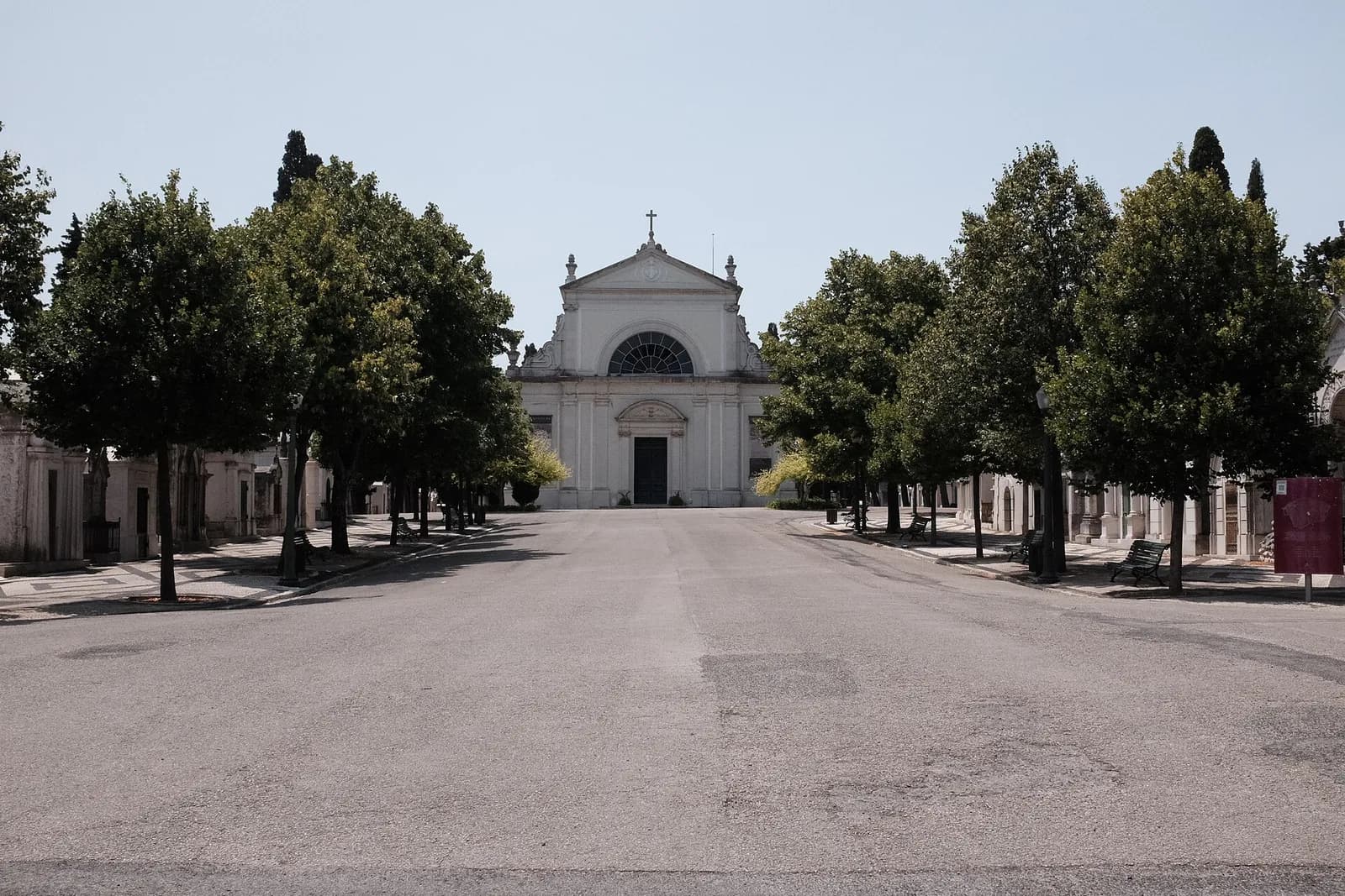 A wide, empty paved avenue lined with trees and rows of small stone mausoleums, leading to a white neoclassical chapel with a semicircular window above its door.