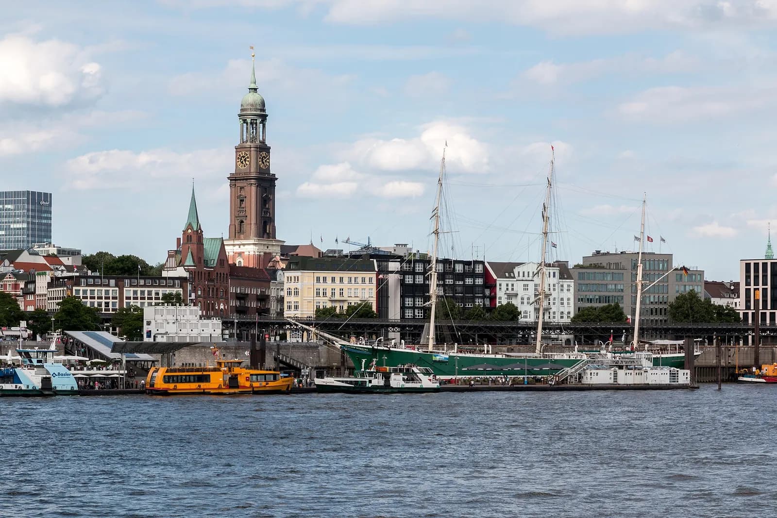 A three-masted sailing ship with a green hull moored on a wide river in front of a city waterfront, with a tall brick church tower with a green copper top behind it and a yellow harbour ferry at the pier.