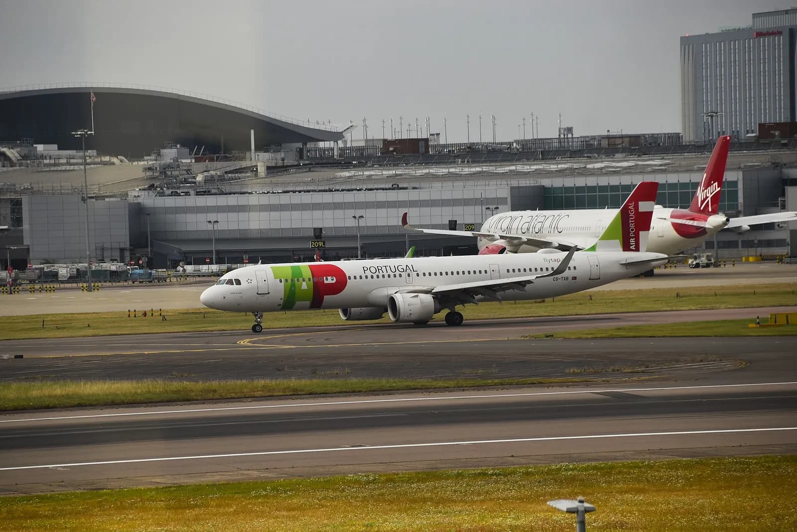 A white airliner with large green and red TAP letters and the word Portugal on its fuselage and a red and green tail, taxiing at a large airport, with a red-tailed Virgin Atlantic jet and terminal buildings behind it.