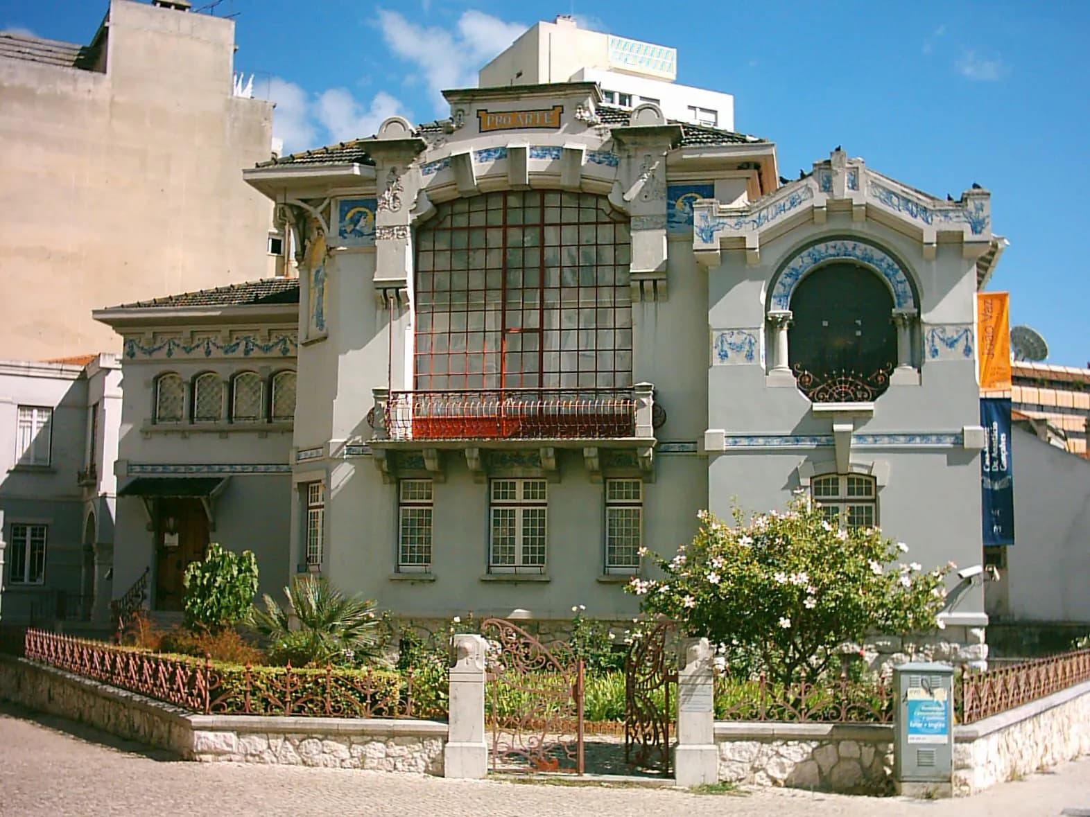 A grey Art Nouveau house with blue and white tile friezes, a tall gridded studio window above a red iron balcony, and a round arched window on the right, behind a low wall with an iron gate and a flowering shrub.