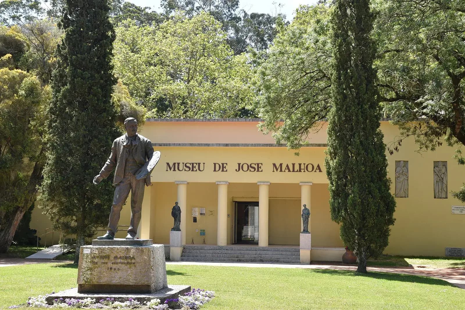 A low pale yellow building with a portico of four columns and the words MUSEU DE JOSE MALHOA across the front, set among tall cypresses and trees. On a stone plinth in the lawn in front stands a bronze statue of a man holding a painter's palette.