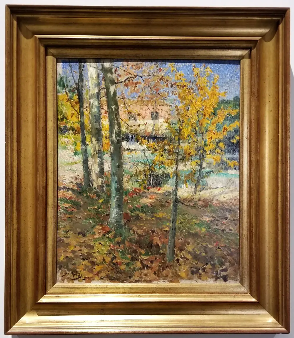 A small framed painting of slender trees with yellow and orange autumn leaves in front of a pale pink house, the ground covered in fallen leaves, all built up from short dabs of bright colour. It hangs in a wide gilt frame on a white wall.