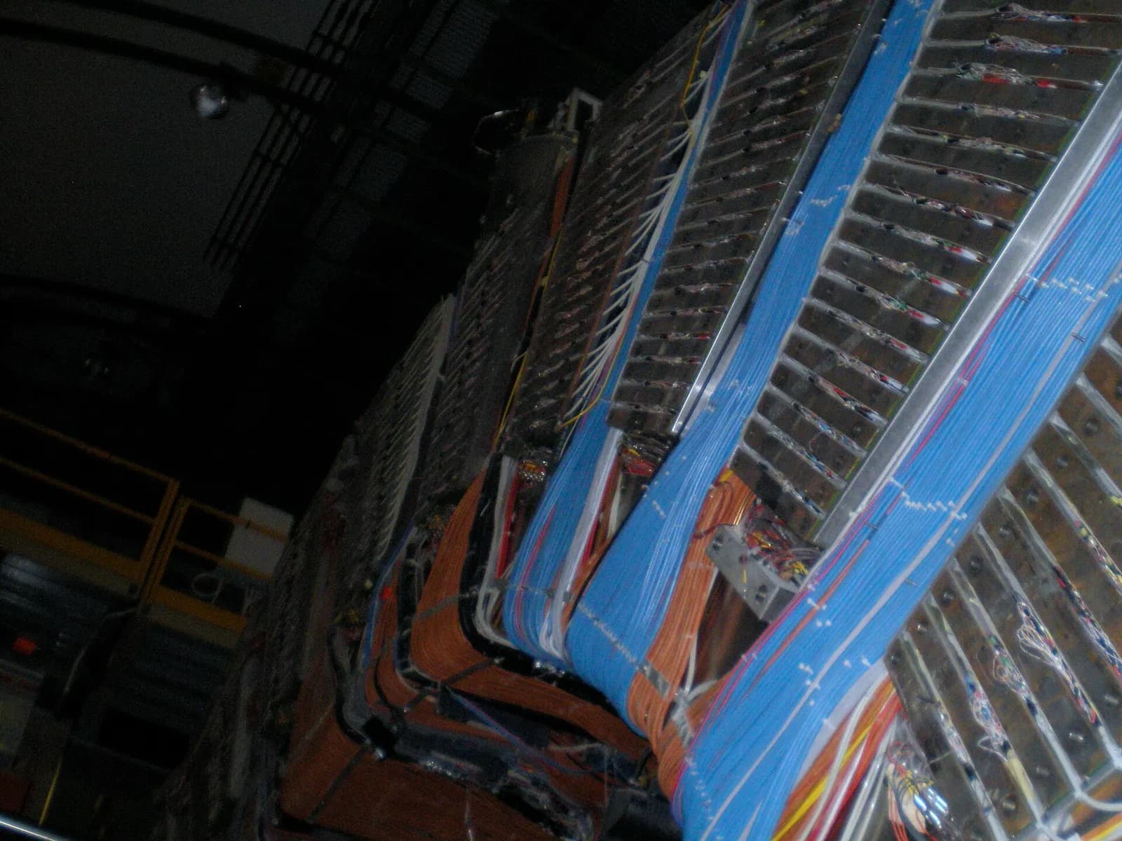 A close view of a huge particle detector seen from below at an angle: stacked rows of grey metal modules bound with thick bundles of blue, orange and red cables, in a dark underground hall.