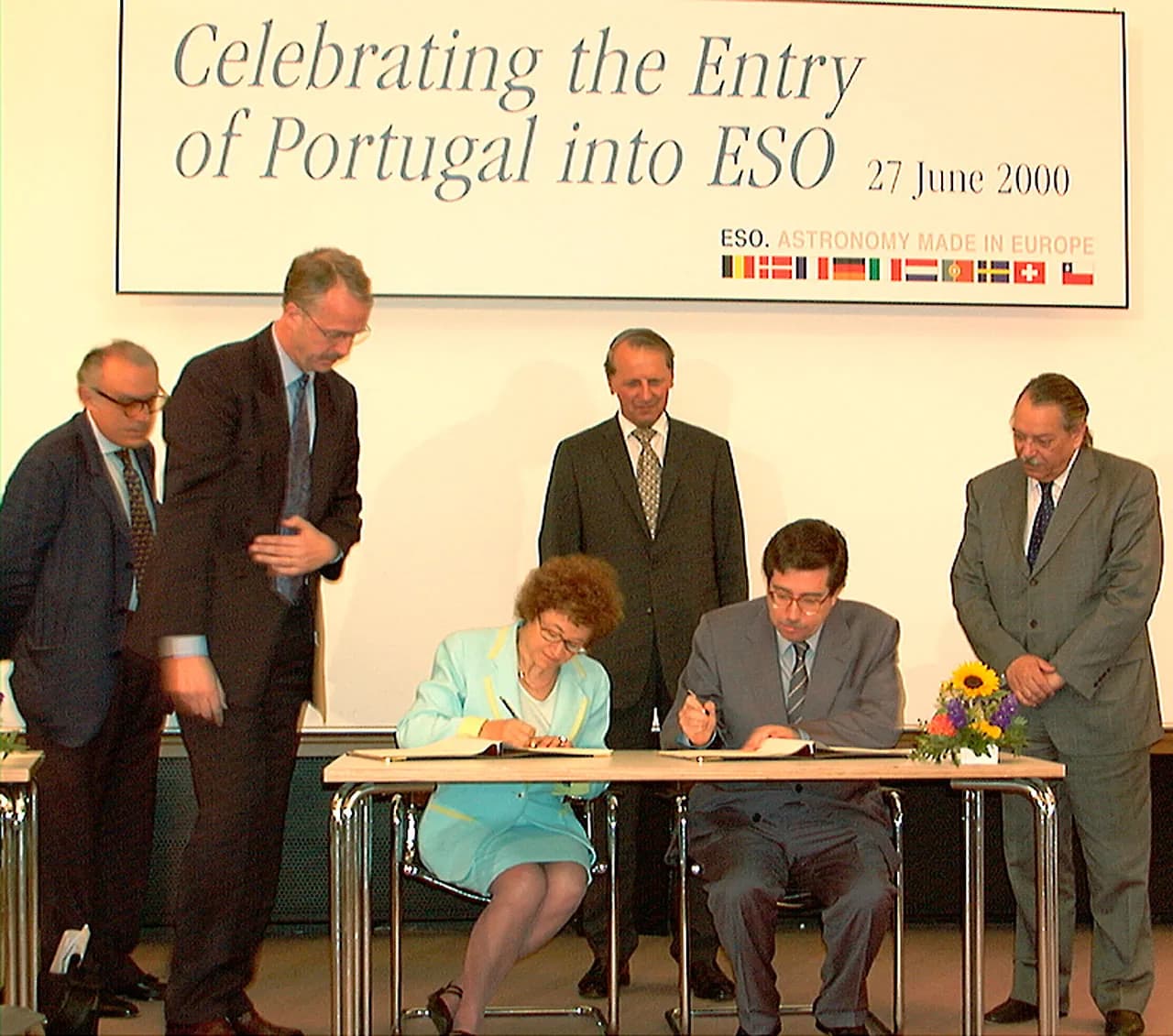 A woman in a pale turquoise suit and a man in a grey suit and glasses sit side by side at a table signing documents, watched by four standing men in suits. A banner above them reads Celebrating the Entry of Portugal into ESO, 27 June 2000, with a row of small national flags.