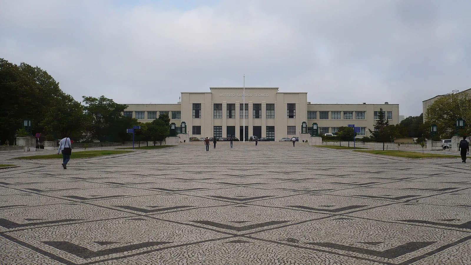 A long, pale, symmetrical modernist building with a taller central block stands at the far end of a wide square paved in black and white stone mosaic, under a cloudy sky. A few people walk across the square.