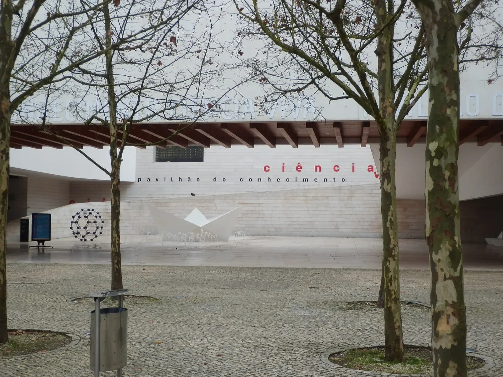 A long white modern building with a deep projecting roof seen across a paved square through bare plane trees. Lettering on its wall reads pavilhão do conhecimento, with ciência in red, and a large model of a carbon molecule and a white folded-paper sculpture stand by the entrance.