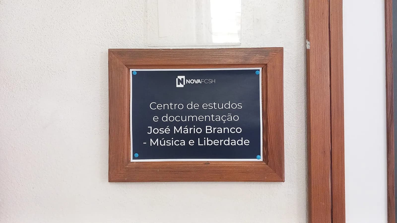 A dark blue sign in a wooden frame on a white wall, with the NOVA FCSH logo and the words Centro de estudos e documentação José Mário Branco – Música e Liberdade.