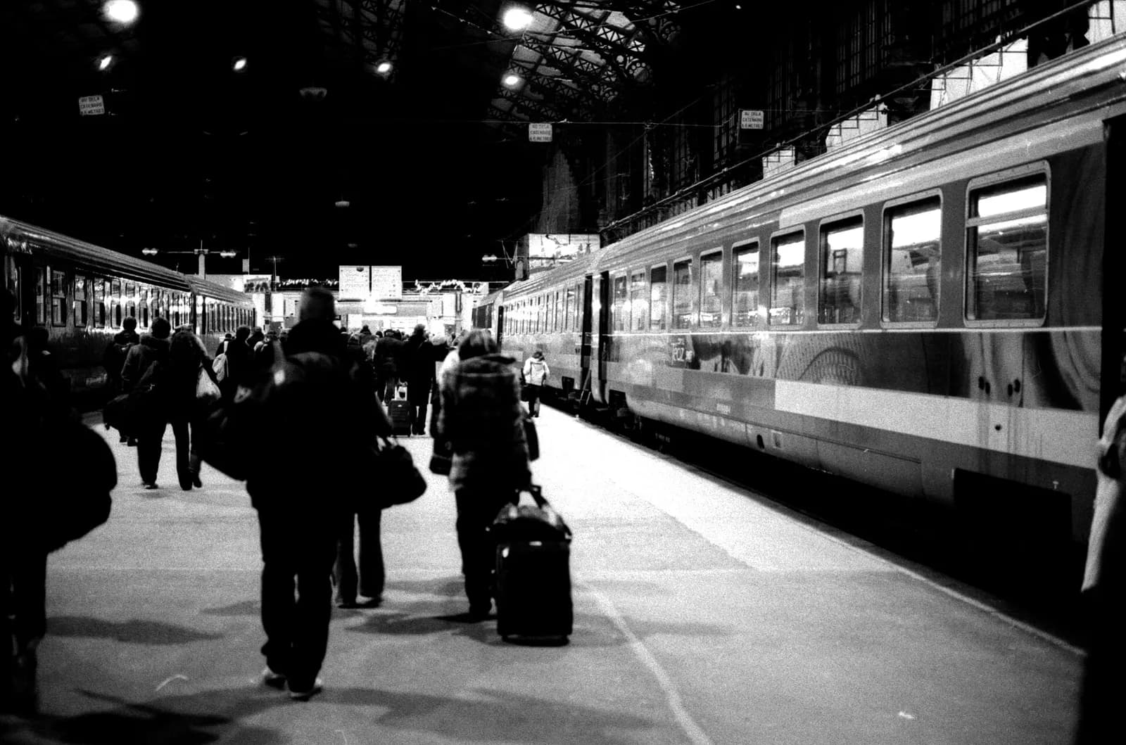 Black and white photograph of a long station platform under an iron and glass roof, with passengers pulling suitcases between two lines of passenger carriages.
