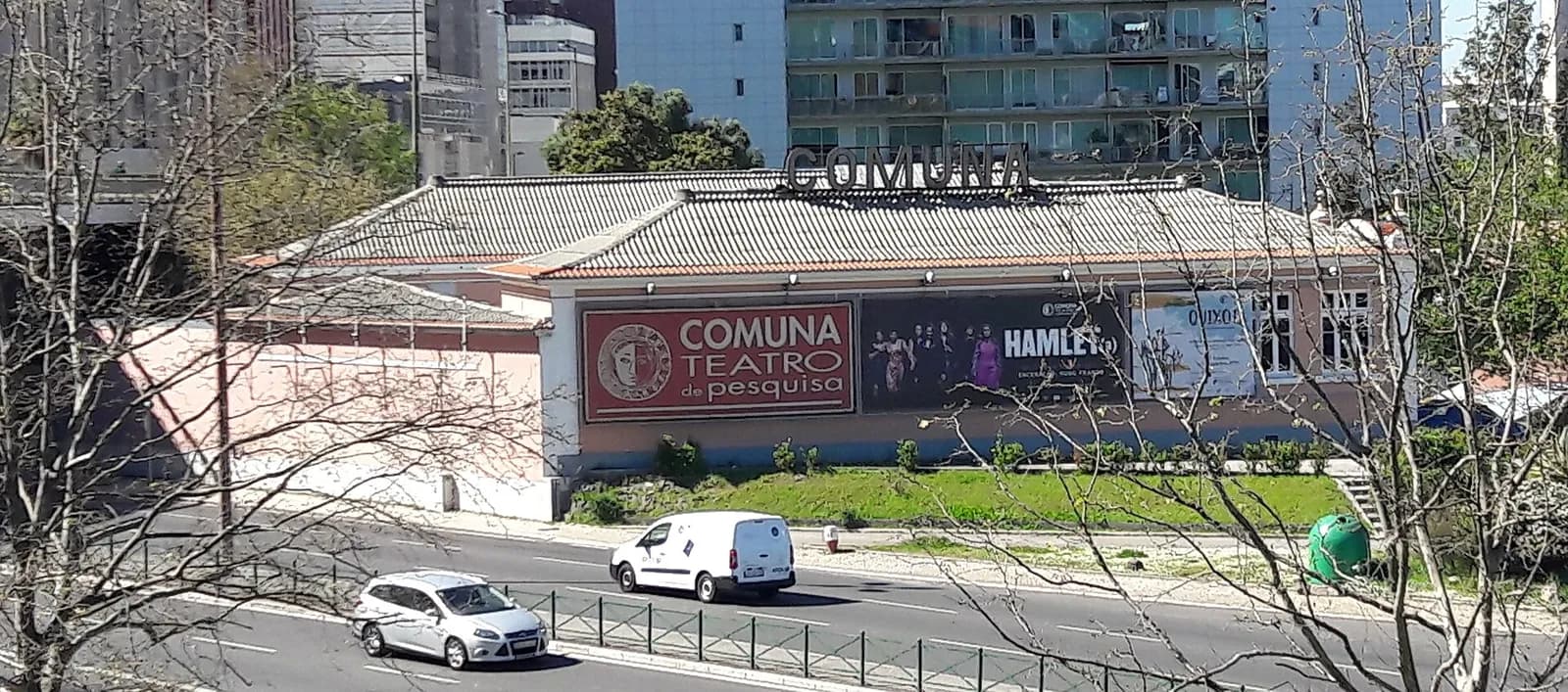 A long, low pink building with a tiled roof beside a busy road, with large posters reading Comuna Teatro de Pesquisa and Hamlet on its front wall, the word Comuna in large letters on the roof and tall modern blocks behind.