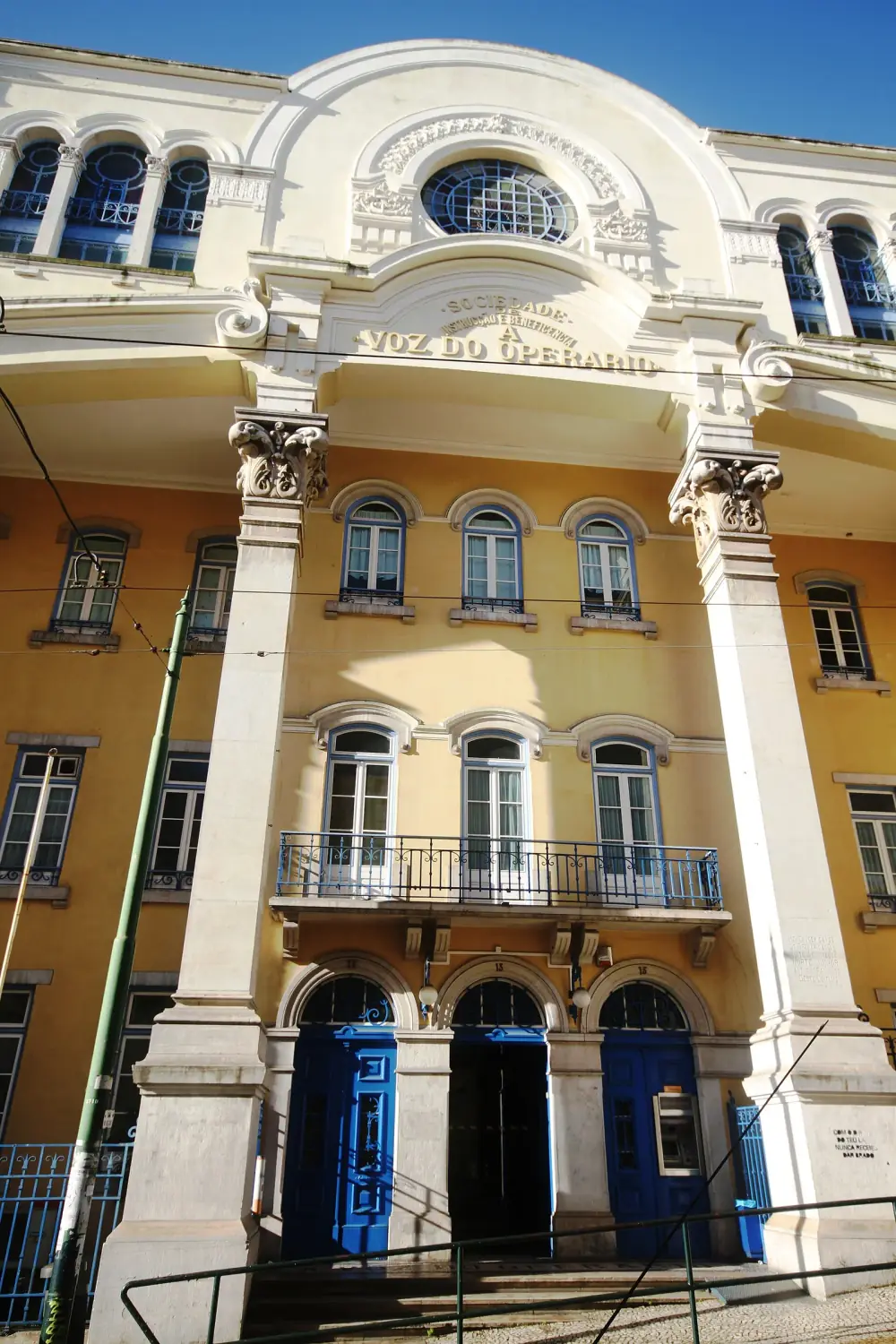 The ornate yellow and white façade of a large building seen from below, with stone columns, a balcony, three arched doorways and the words A Voz do Operário carved above.