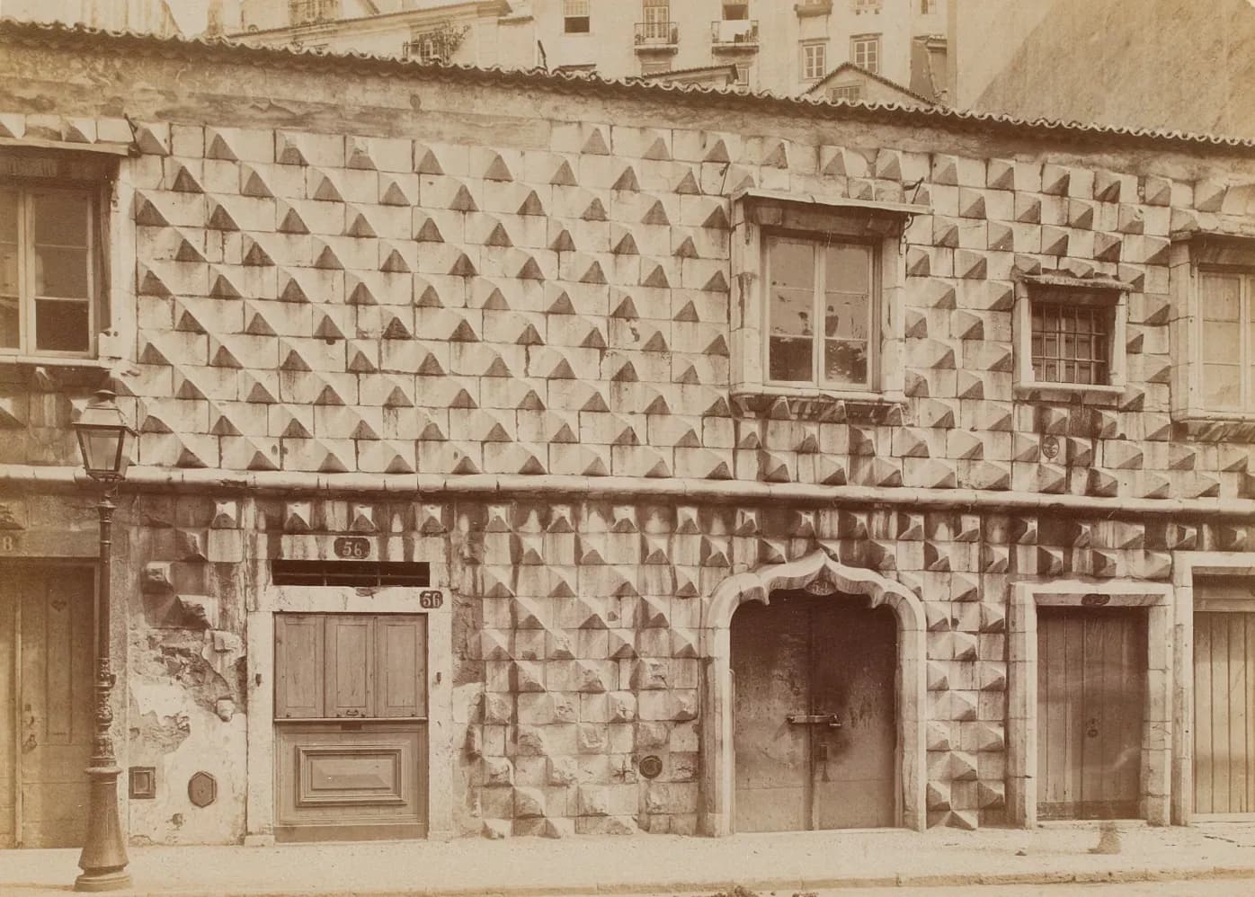 Old sepia photograph of a two-storey street front faced with rows of pyramid-shaped stones, with a pointed stone doorway, several wooden doors, a few windows, a gas lamp post on the pavement and a plain tiled roof just above the second floor.