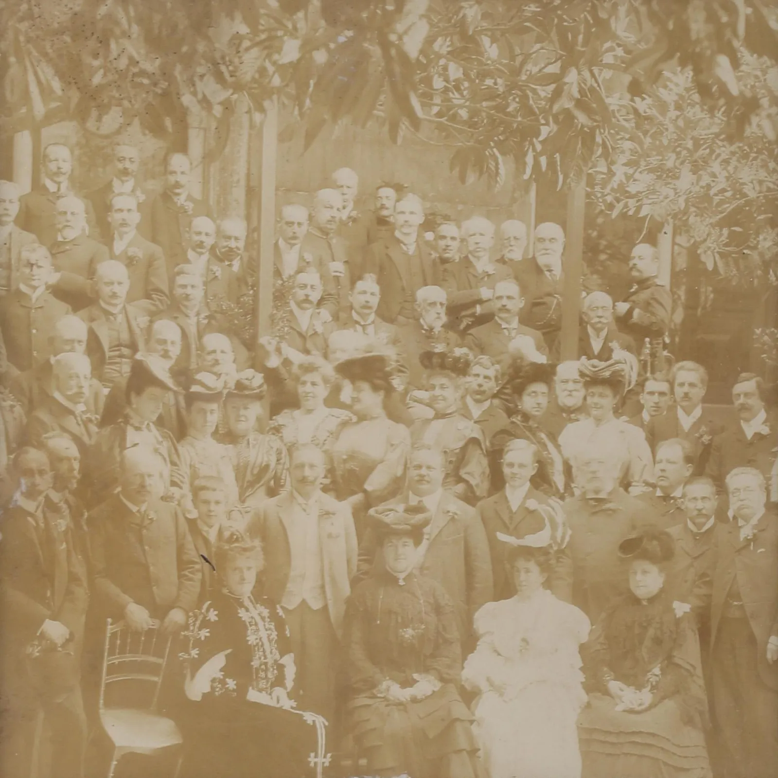 A sepia group photograph of several dozen people under trees: four women in hats seated in the front row, men in dark suits, one man in a pale suit and two teenage boys standing behind them, more women in large hats, and rows of older men in suits at the back.