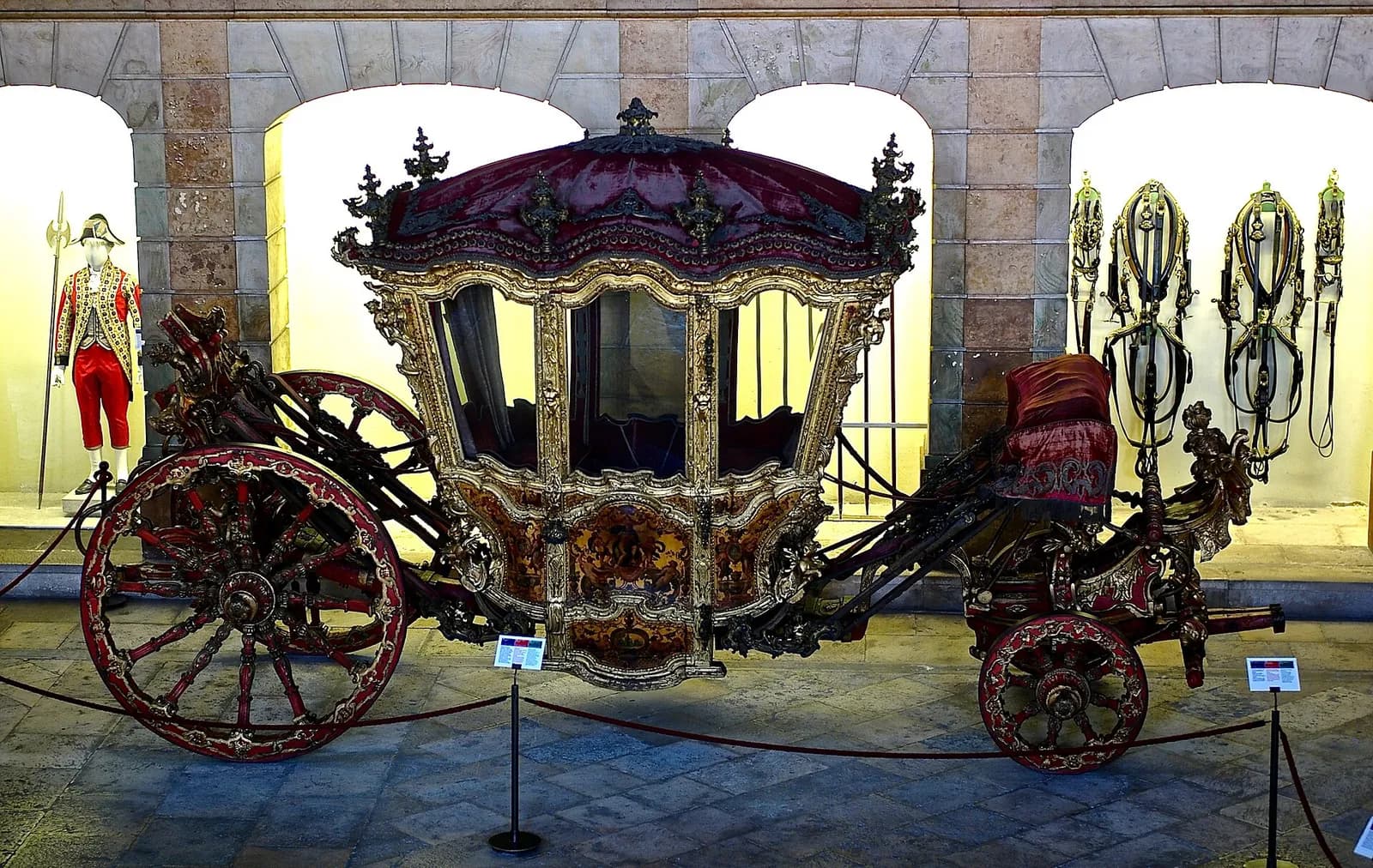 A gilded and carved eighteenth-century coach with a red velvet roof, glass windows, painted panels and large red and gold wheels, displayed behind a rope under stone arches, with a footman's livery on a mannequin to the left.