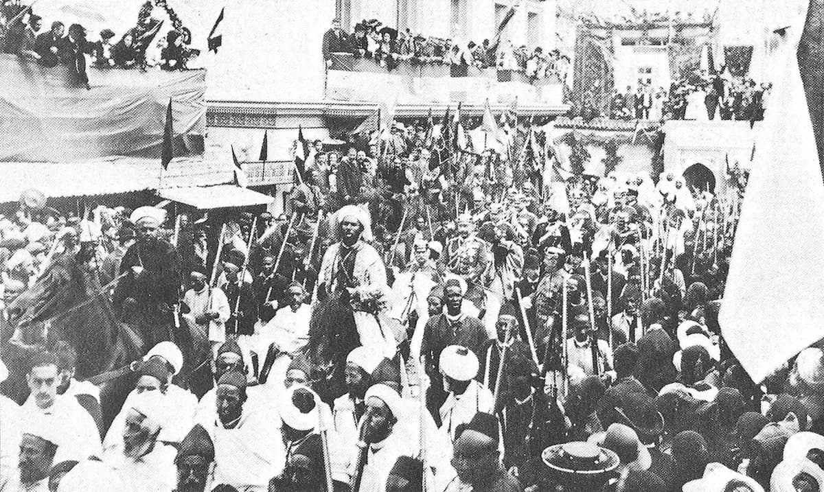 A dense crowd of men in turbans and white robes fills a street hung with flags; men on foot and on horseback carrying long lances move through it, and spectators watch from balconies and rooftops.