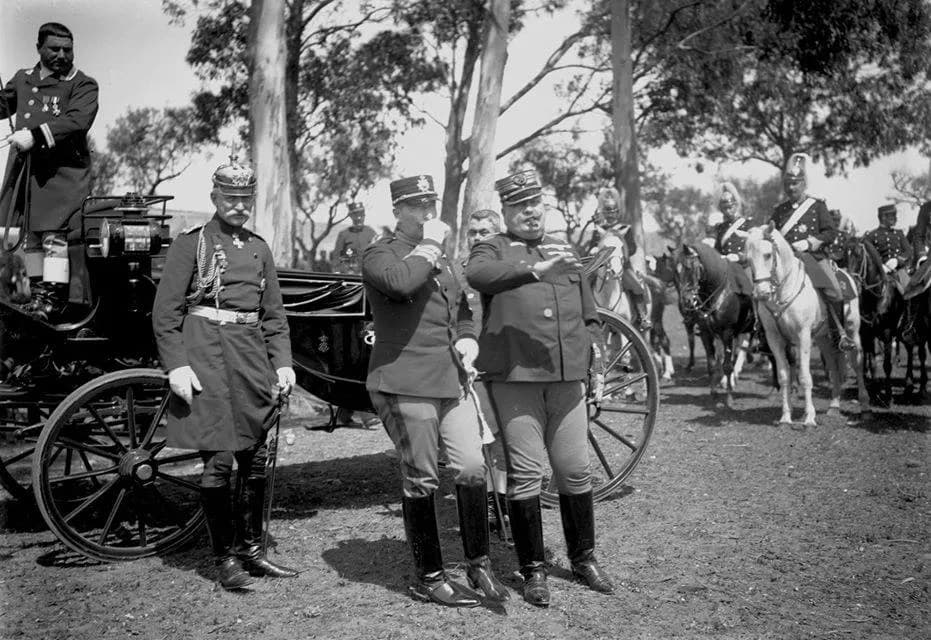 Officers in uniform stand on bare ground under eucalyptus trees beside an open carriage. At the centre two men in Portuguese army caps and high boots talk, one stout and smoking a cigar, the other raising a white-gloved hand to his face; at left an officer wears a spiked helmet. Cavalrymen in plumed helmets sit on horses behind them.