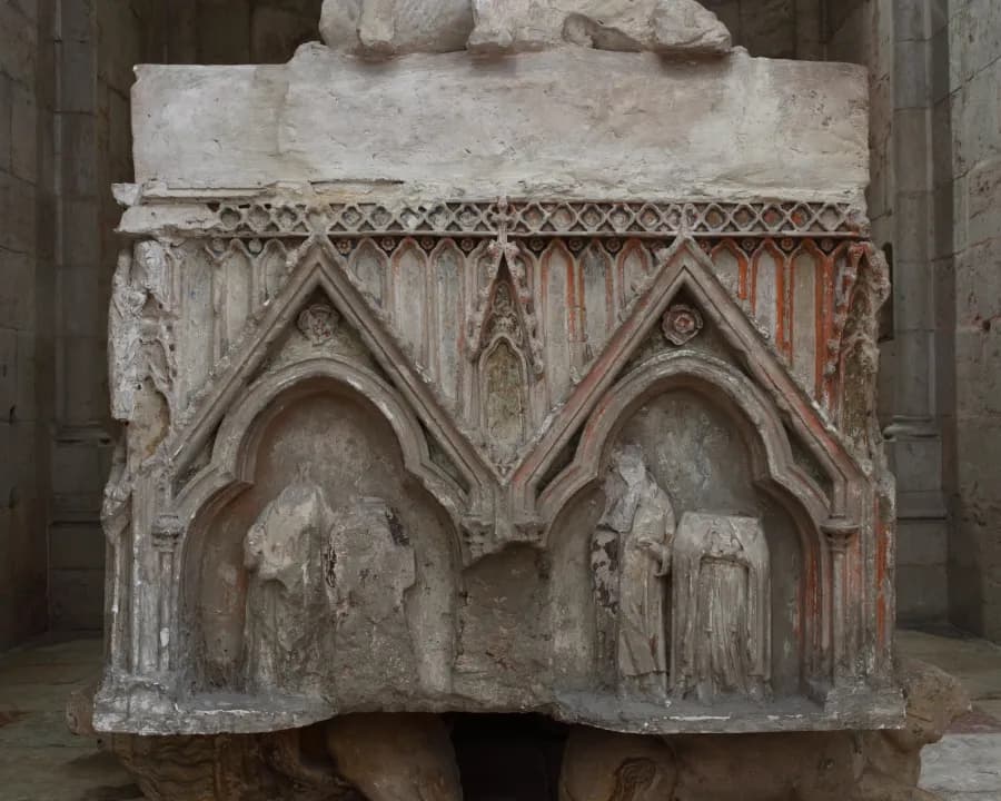 Part of a stone tomb chest carved with two pointed Gothic arches under gables, each sheltering worn figures, with traces of red paint, resting on carved supports in a bare stone church.