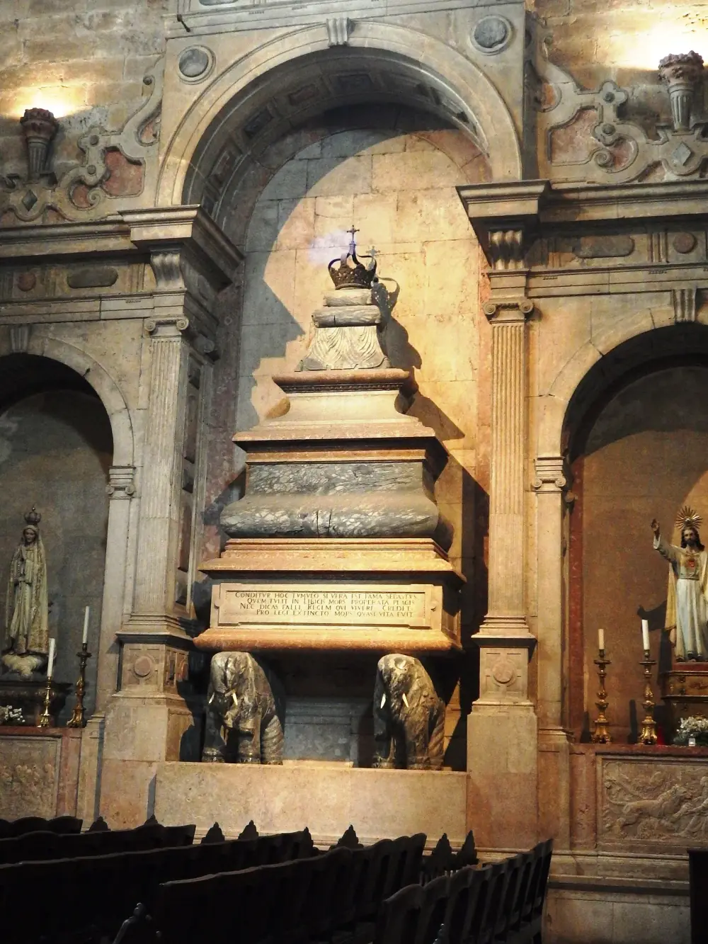 A tall marble tomb under a stone arch in a church, topped by a crown and resting on two carved elephants with ivory tusks, with a Latin inscription on its base. Statues of the Virgin and of Christ stand in side niches.