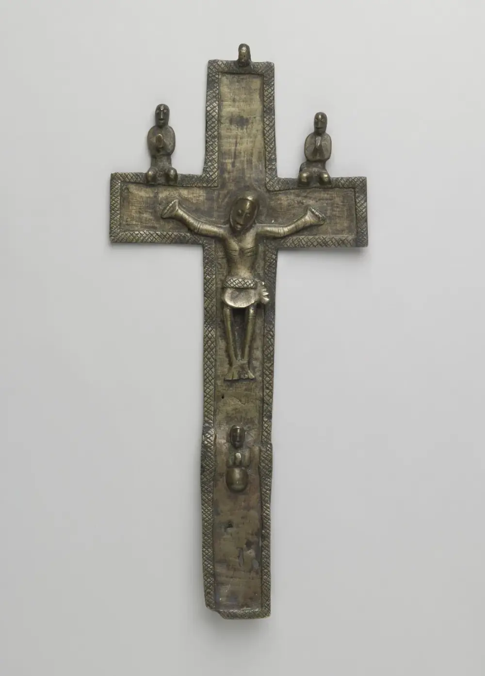 A cast copper-alloy crucifix with a hatched border: Christ with outstretched arms in the centre, two small kneeling figures with hands joined on the crossbar and a third below his feet.