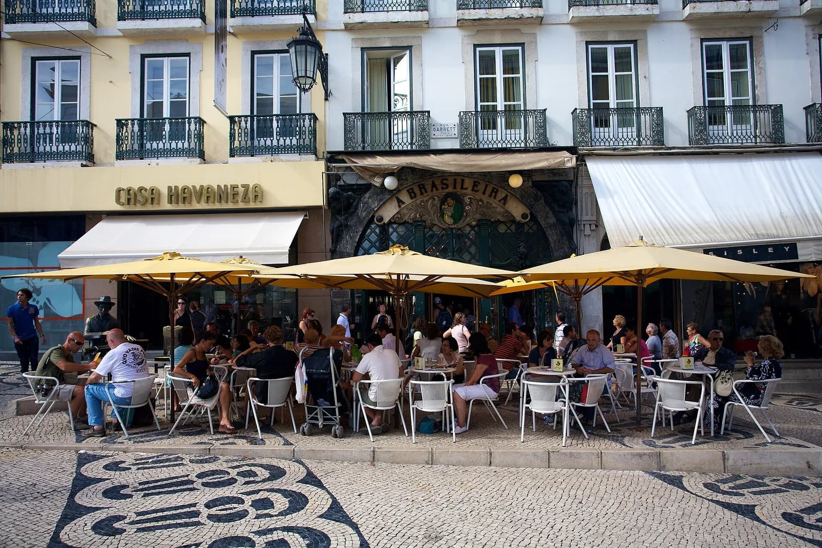 A café terrace crowded with people at white tables under yellow umbrellas, in front of an ornate green shopfront with the gilt sign A Brasileira. At the far left, beside the Casa Havaneza shop, a dark bronze figure in a hat sits at a small table among the customers.