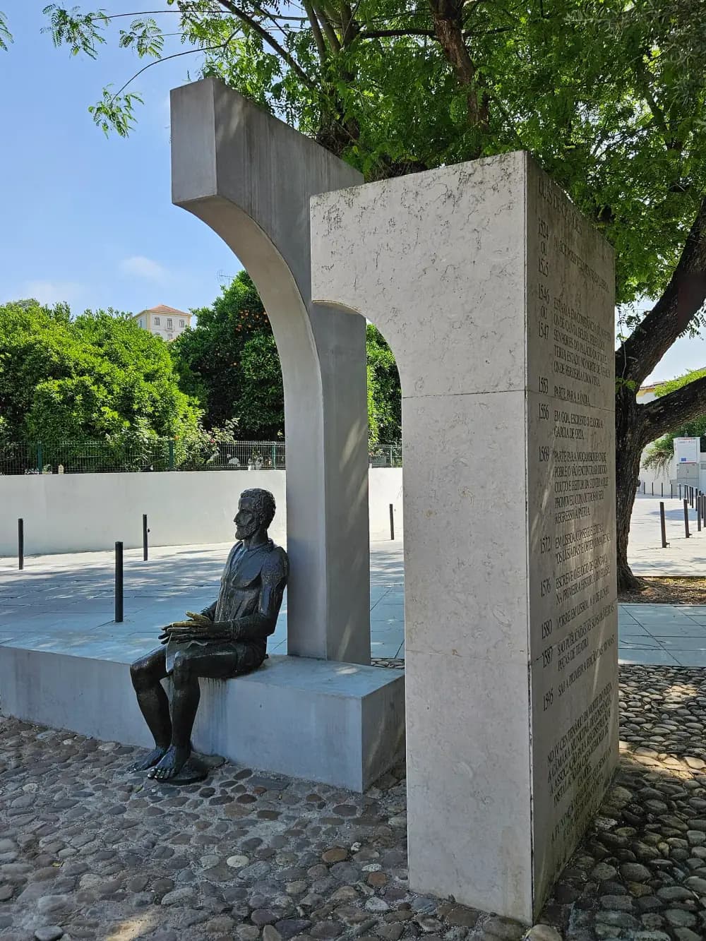 A bronze of a bearded, barefoot man sits on a long stone bench, hands resting in his lap, in front of two tall curved stone slabs that form an arch. Beside him stands a stone slab carved with dates and lines of text, with trees and a paved square behind.