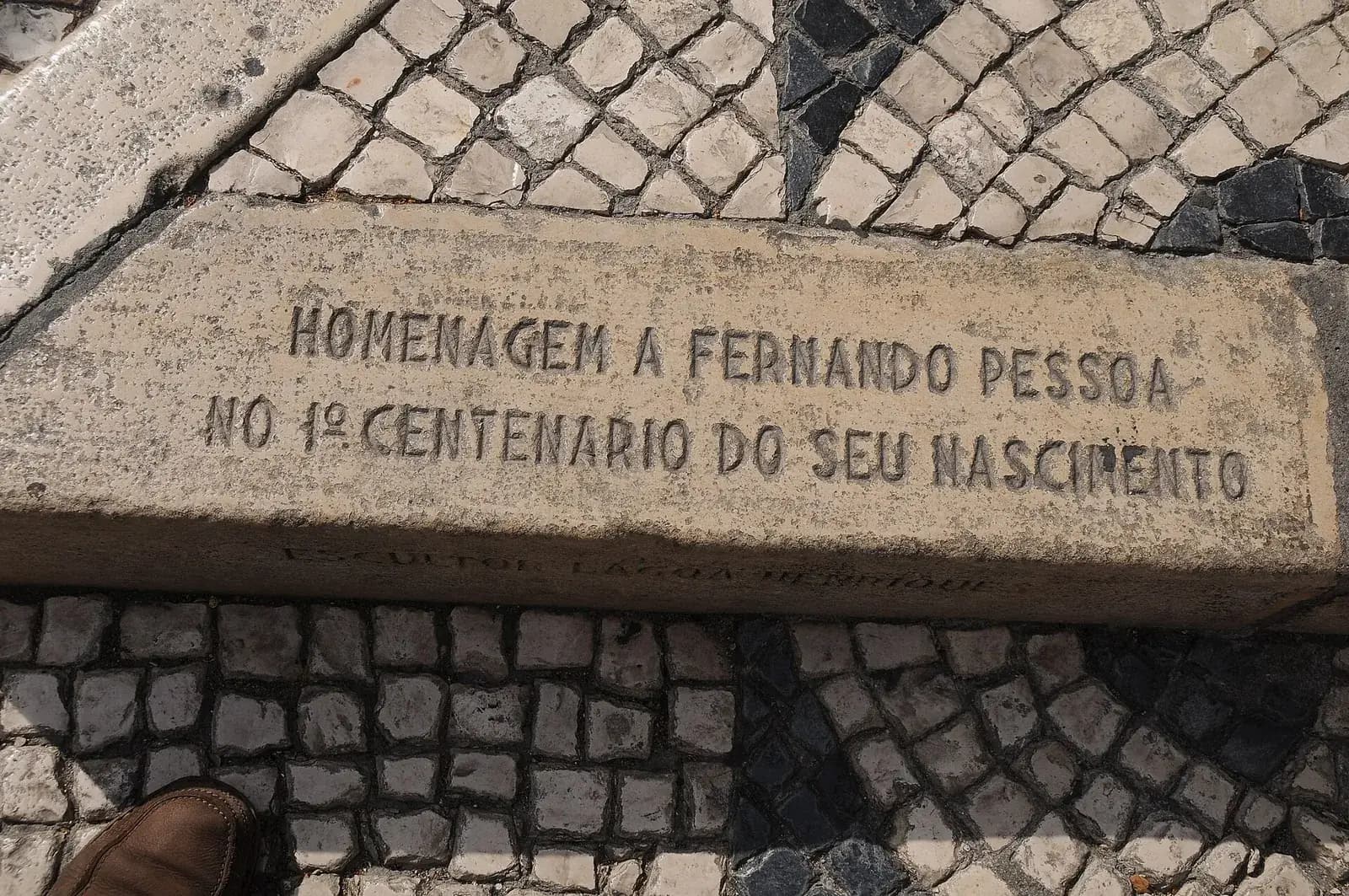 A worn stone kerb set into a black-and-white cobbled pavement, carved in capitals: Homenagem a Fernando Pessoa no 1º centenario do seu nascimento. A fainter line below reads Escultor Lagoa Henriques. A visitor's brown shoe is just visible at the bottom left.