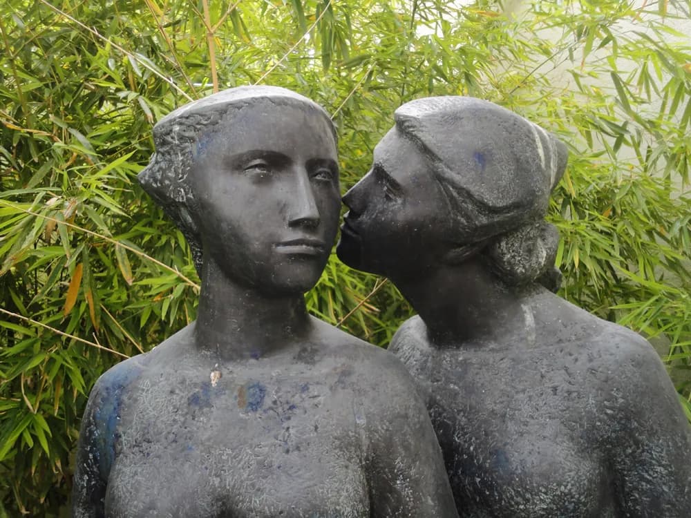Close view of two bronze heads and shoulders of young women against bamboo leaves. The one on the right leans in with her lips close to the cheek of the other, who looks straight ahead.
