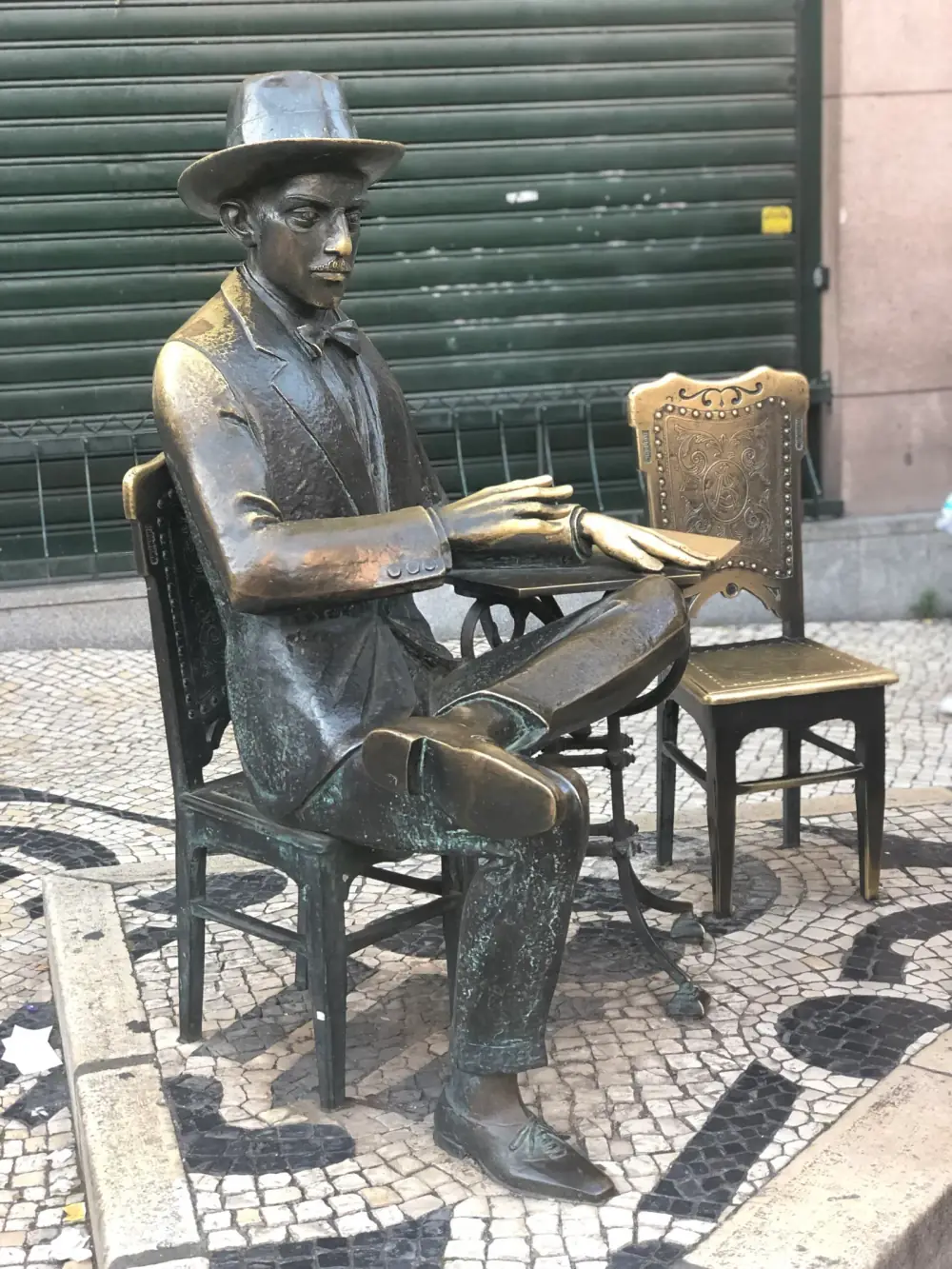 A bronze statue of a thin, moustached man in a hat, bow tie and three-piece suit sits cross-legged on a bronze chair at a small bronze café table, his hands resting on it. Across the table stands an empty bronze chair with a carved back, on black-and-white cobbles in front of a closed green shutter.