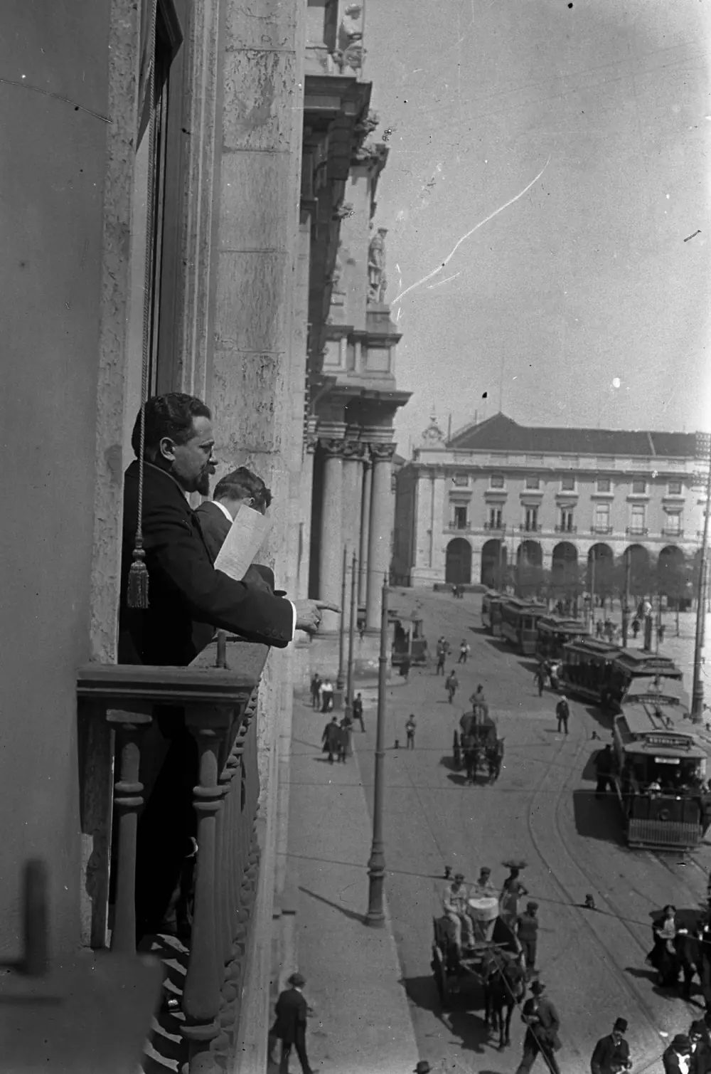 A bearded man in a dark coat leans over a wooden balcony on the upper floor of a stone building, pointing down at a busy square. Beside him a younger man holds a sheet of paper. Below are trams, horse-drawn carts and pedestrians, with an arcaded building in the background.