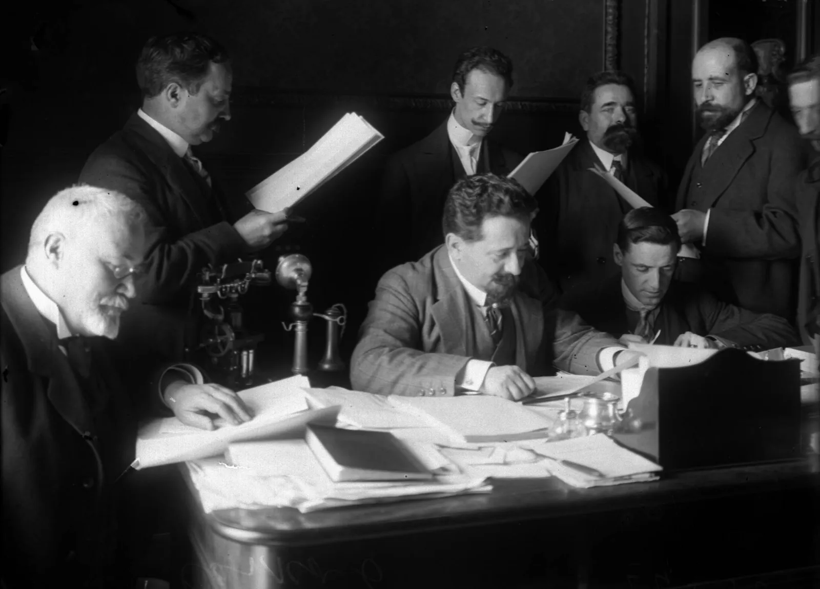 A bearded man in a light suit sits at a desk covered in papers, looking down at them, beside an older white-haired man reading and a younger man writing. Five men in dark suits stand behind them, several reading sheets of paper. A candlestick telephone stands on the desk.