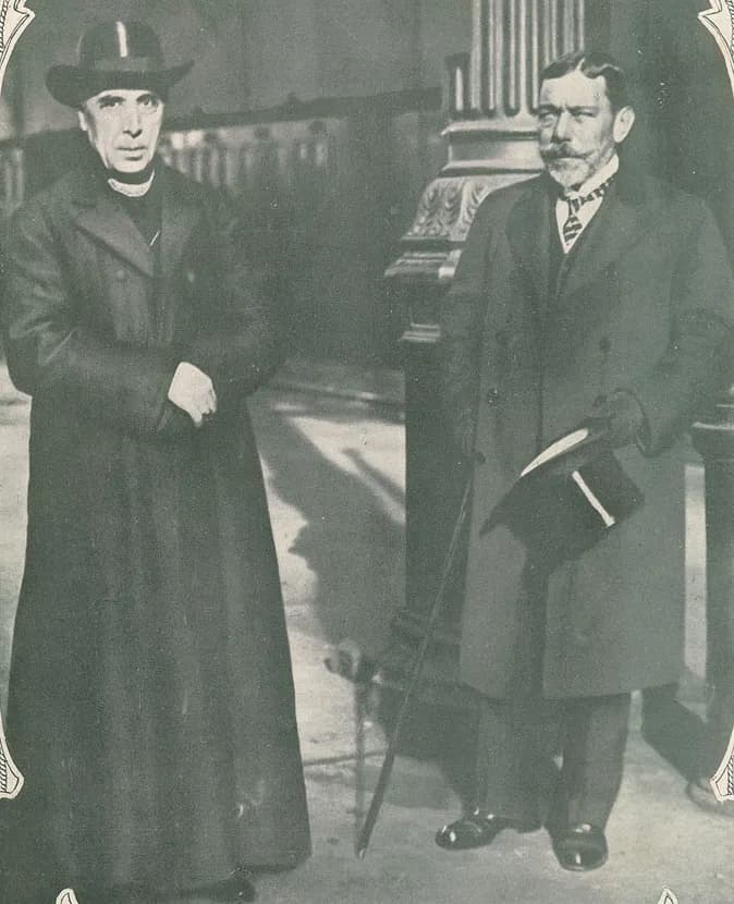 Two elderly men stand side by side outdoors in front of a stone column. On the left a clean-shaven cleric in a long black coat and a wide-brimmed hat holds his hands together; on the right a bearded man in an overcoat holds a top hat and a cane.